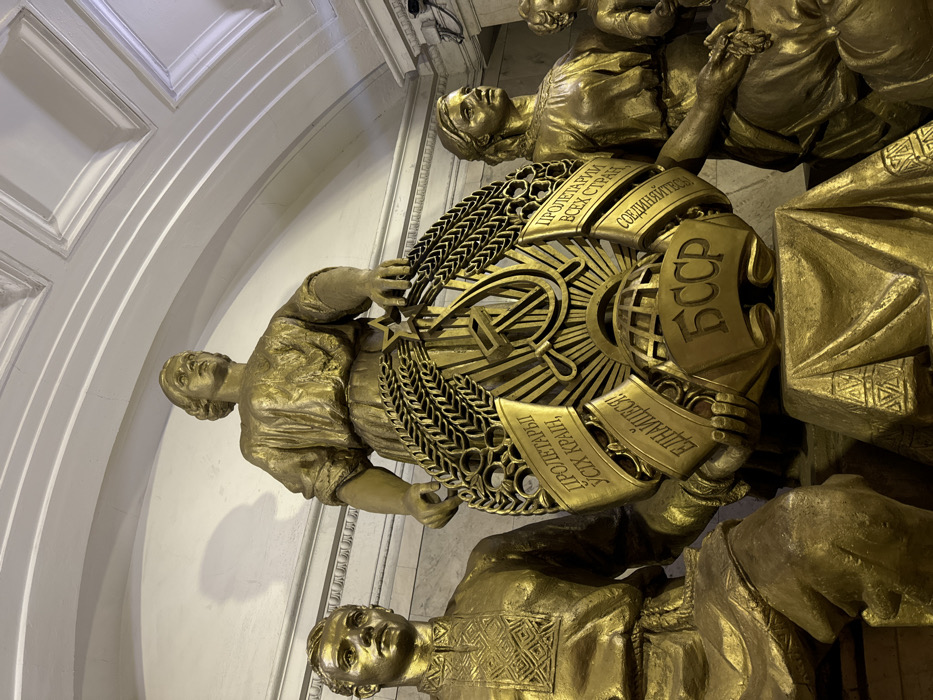 The image displays a detailed view of a sculptural group located within the Kievskaya Station (Koltsevaya Line) of the Moscow Metro, Moscow, Russia. The scene features three prominent, gold-colored statues of idealized human figures positioned against a light-colored, classically adorned wall beneath an arched, coffered ceiling.
In the center, a standing female figure in a flowing garment holds a large, circular golden emblem. This emblem prominently features the Soviet hammer and sickle symbol, radiating sun rays, positioned above a globe, all encircled by a wreath of wheat ears with a red star at its apex. Banners unfurling from the emblem bear the text "ПРОЛЕТАРІЇ УСІХ КРАЇН, ЄДНАЙТЕСЯ!" (Ukrainian for "Proletarians of all countries, unite!"), "ПРОЛЕТАРИИ ВСЕХ СТРАН, СОЕДИНЯЙТЕСЬ!" (Russian for "Proletarians of all countries, unite!"), and "БССР" (Byelorussian Soviet Socialist Republic).
To the left of the central figure, a seated male statue is depicted in traditional embroidered attire. To the right, a seated female statue in traditional dress is shown with a partially visible child figure next to her. All figures exhibit composed expressions. The central emblem rests on a golden pedestal draped with stylized fabric. The entire composition is rendered in a Socialist Realist style, embodying themes of Soviet unity and the friendship of peoples.
