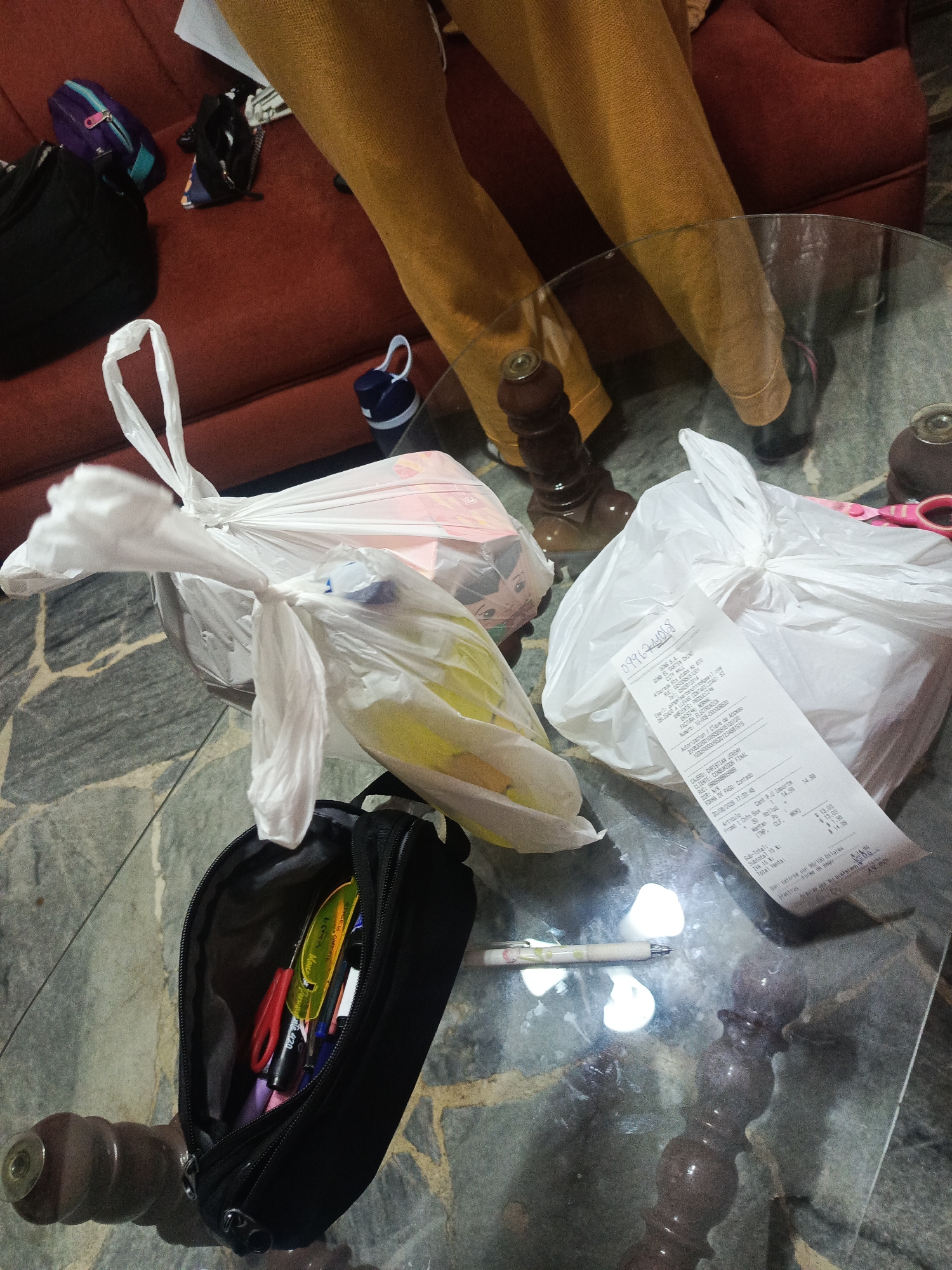 The image shows a top-down view of a glass coffee table with several items on it. In the background, a person is sitting on a reddish-brown couch, wearing mustard-colored pants. On the couch, to the left of the person, are a small purple and teal bag and a black bag.
On the coffee table, there are two white plastic grocery bags, one tied shut and the other partially open, revealing yellow items inside. To the right of the grocery bags is a receipt with Spanish text. In front of the grocery bags is an open black pencil case containing pens and scissors. To the left of the pencil case is a white pen with a floral design. The coffee table has a dark, patterned surface. The legs of the coffee table appear to be made of dark wood with decorative carvings.
Based on the receipt visible in the image, which displays "BODEGA SANTA ELENA C.C. EL DORADO" and other details, this scene is located in Guayaquil, Ecuador. The date on the receipt, "20/06/2020", is also visible.