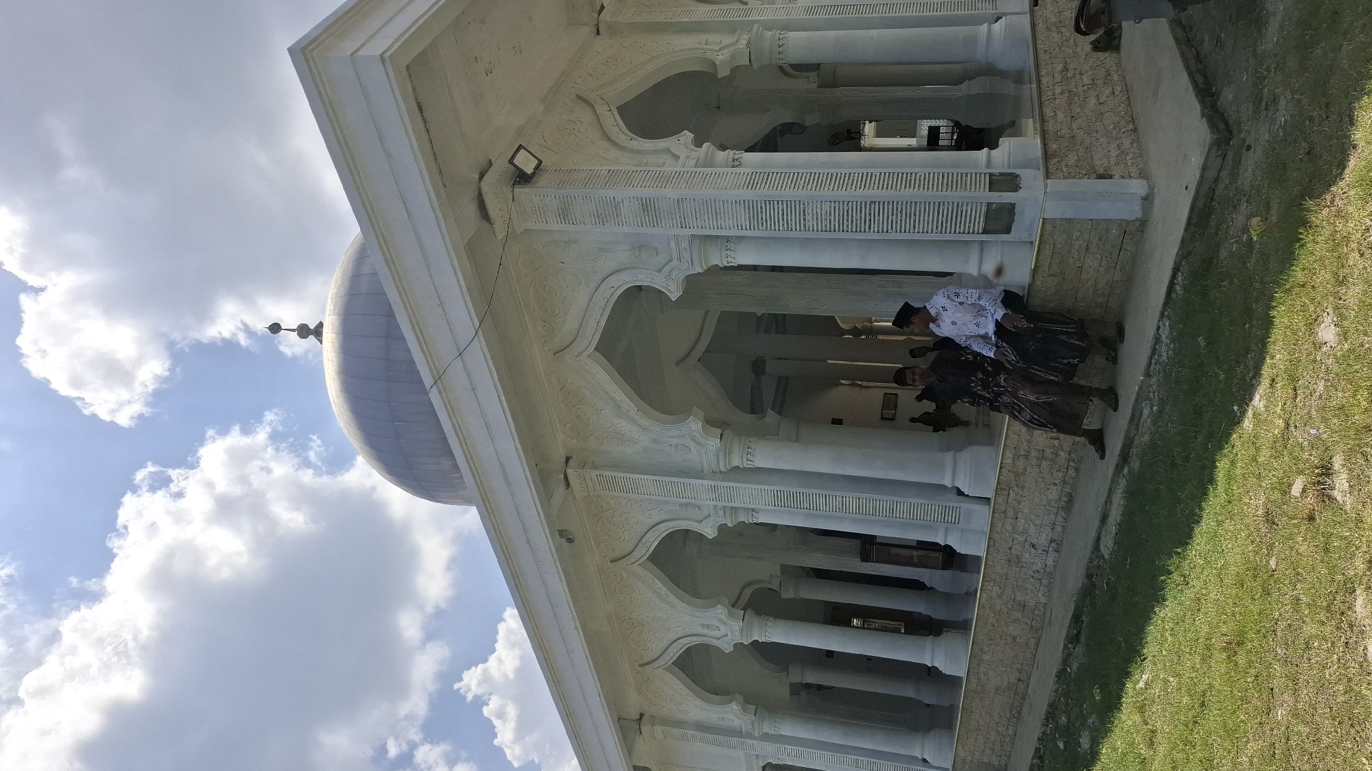 Two men are sitting on a low stone wall in front of a white mosque in Grong-grong, Indonesia. The man on the left is wearing a dark brown patterned sarong and a dark shirt. The man on the right is wearing a black sarong and a white shirt with black embroidered patterns and a black cap. The mosque has a dome and is decorated with intricate carvings around its arches. The sky is blue with scattered white clouds. The ground in front of the mosque is covered with green grass.