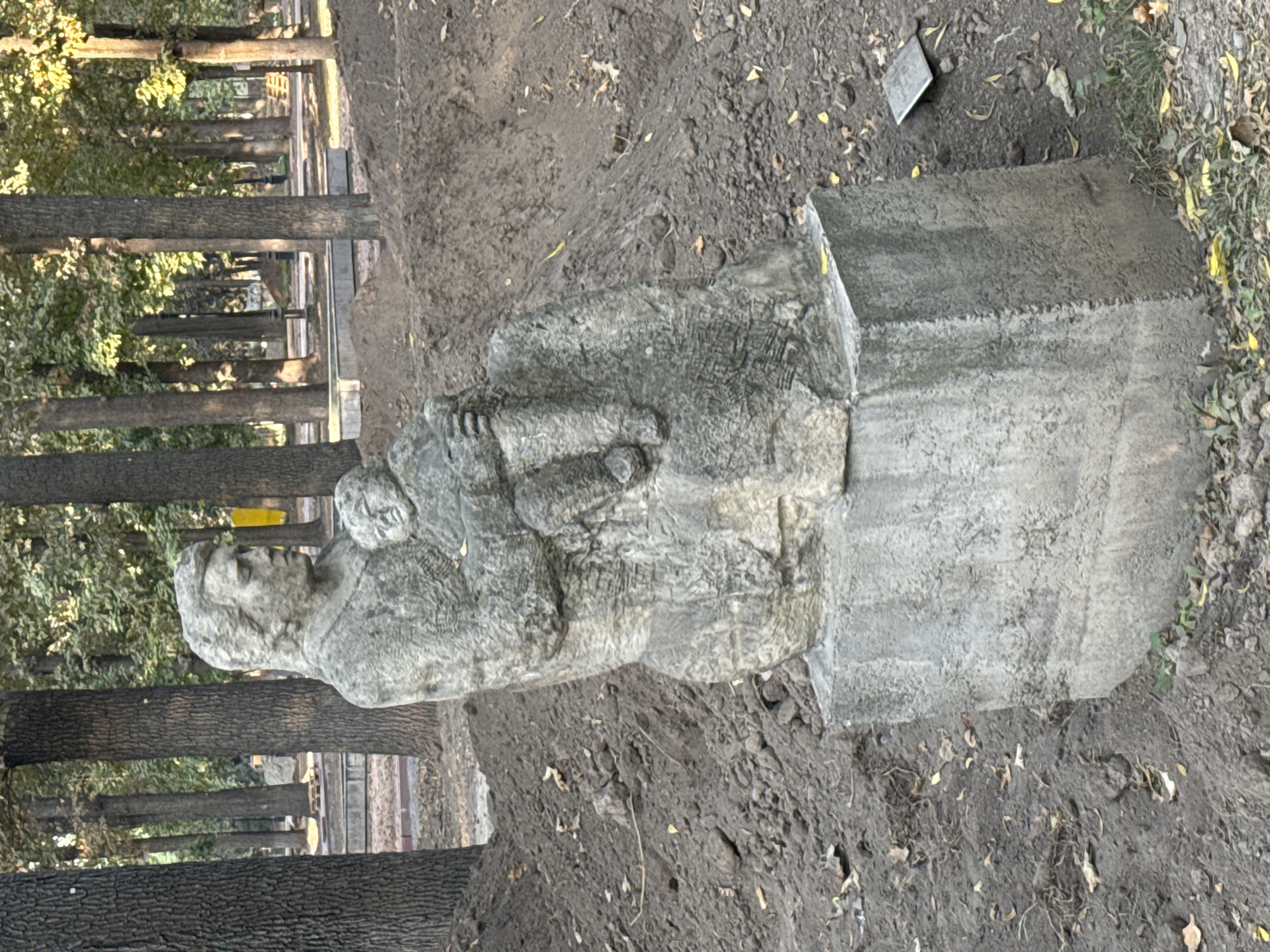 A weathered stone sculpture of a woman holding a child sits on a concrete pedestal, surrounded by disturbed earth and autumn leaves in a park. The sculpture depicts a seated woman with her child cradled in her arms, both figures rendered with a rough, textured finish. The woman's head is turned to the side, and her facial features are indistinct due to the style of the carving. The child appears to be an infant, nestled against the woman's chest.
The pedestal is a simple, rectangular block of concrete, showing signs of age and weathering. The ground around the sculpture is uneven, with piles of dark soil suggesting recent excavation or landscaping. Scattered brown and yellow leaves are visible on the soil and at the base of the pedestal, indicating it is likely autumn.
In the background, tall trees with bare branches and some remaining green foliage form a dense forest. The sunlight filters through the trees, creating dappled shadows on the ground. Further in the background, elements of a park or urban setting can be discerned, including what appears to be a pathway and some man-made structures, possibly benches or planters, partially obscured by the trees. The overall impression is of a public outdoor space, likely a park in Bishkek, Kyrgyzstan, given the context provided, undergoing some form of maintenance or preparation.