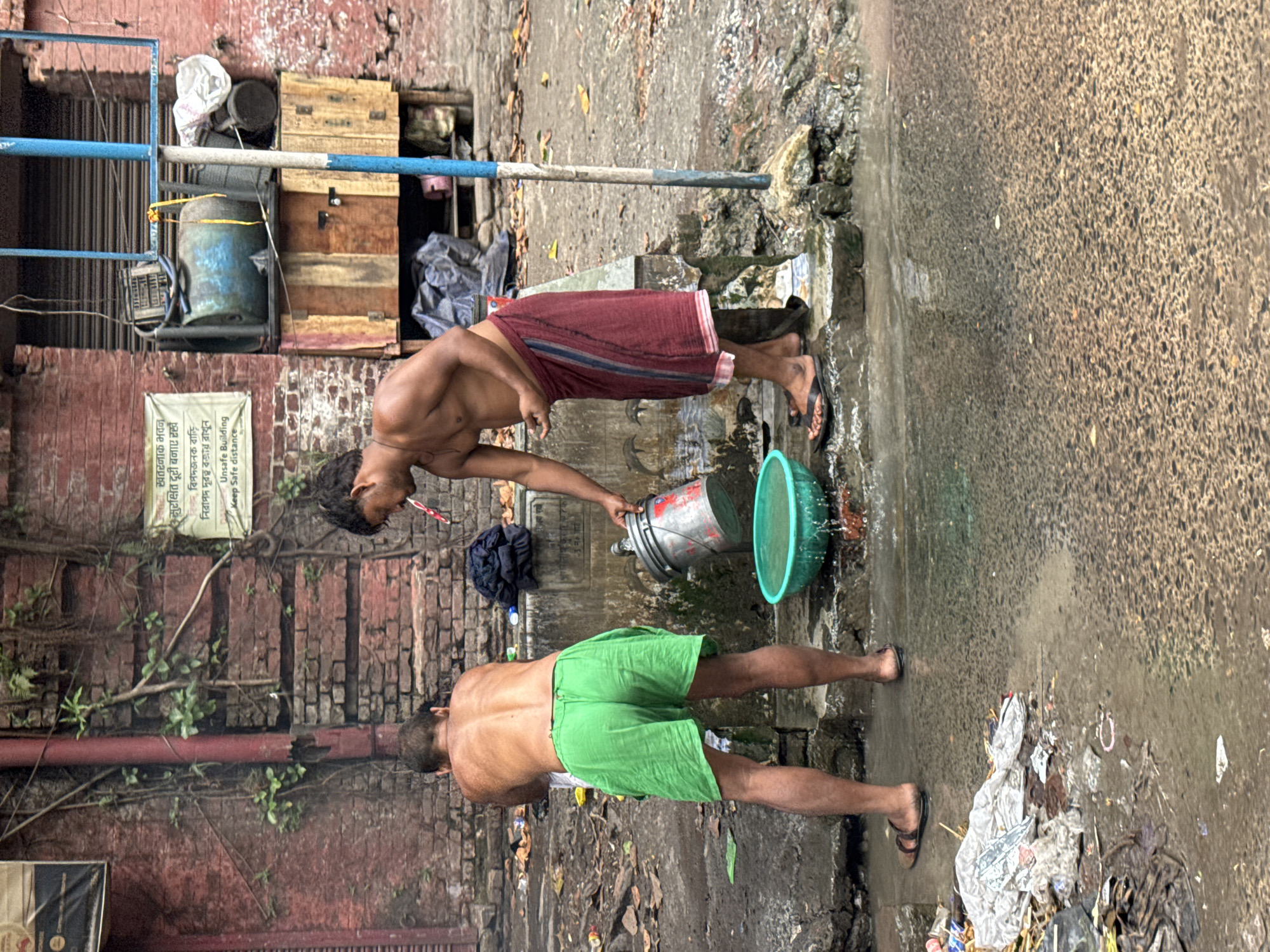 This image, set in Kolkata, India, captures an outdoor scene likely in an urban alleyway or street during daytime. Two shirtless men are engaged in washing activities. The man on the left wears a dark red lungi and is holding a toothbrush in his mouth, appearing to pour water from a silver metal bucket into a bright green plastic basin on the ground. He faces towards the right side of the frame. The man on the right wears green shorts and is bent over, facing away from the viewer. Both men are wearing flip-flops.
The ground around them is wet and appears dirty, with various debris and litter scattered. On the left side of the frame, a reddish-brown brick wall with some climbing plants is visible. A white sign is affixed to this wall, bearing text in Bengali and English that reads: "খতরনাক ভবন সুরক্ষিত দূরত্ব বজায় রাখুন বিপদজনক বাড়ি নিরাপদ দূরত্ব বজায় রাখুন Unsafe Building Keep Safe distance," warning about a dangerous building. Above the wall, a blue metal railing structure is present, along with what appear to be wooden crates and a blue barrel. The overall scene depicts a common, daily ablution routine unfolding in a bustling and somewhat gritty urban environment.