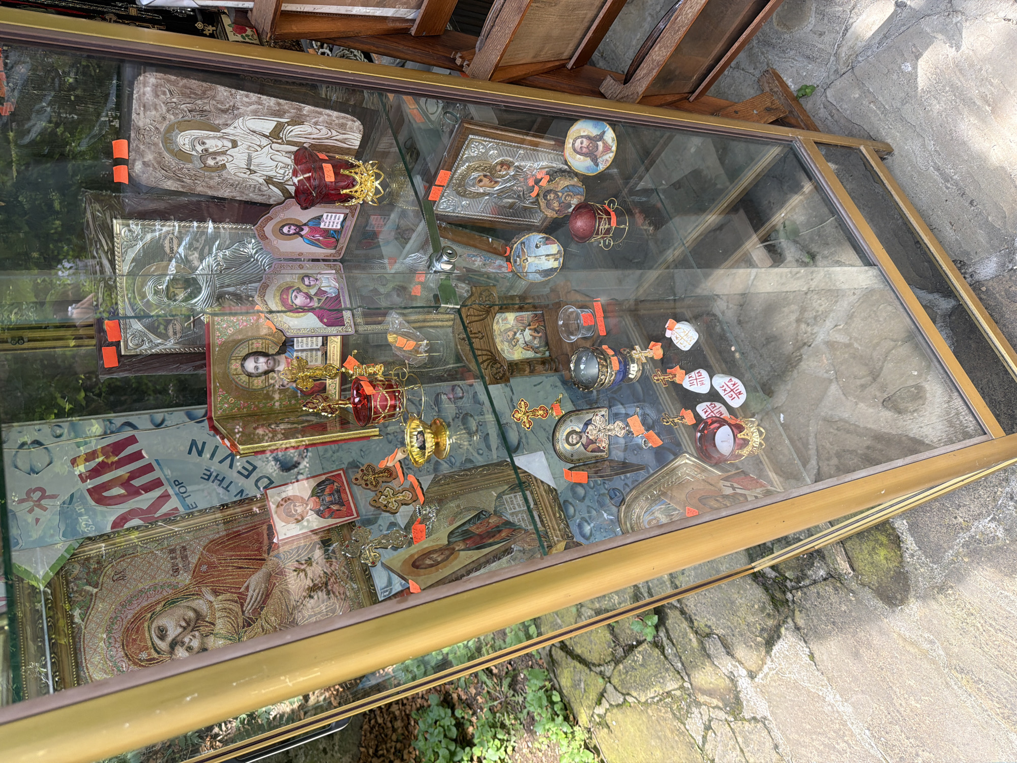 The image displays a vertically oriented, gold-framed glass display case, positioned outdoors on a paved stone or concrete surface in Basarbovo, Bulgaria. The case contains multiple shelves showcasing various Eastern Orthodox religious items. Prominent objects include numerous icons, such as a large top-shelf icon of the Virgin Mary holding the Child Jesus, intricate silver-toned relief icons, and smaller icons depicting Christ, the Virgin Mary, and the Crucifixion. Additionally, gold-colored ornamental crosses, red and clear glass candle holders, and a small gold bowl are visible. Small orange rectangular tags, likely price labels, are affixed to several items. Reflections on the glass case show overhead foliage, trees, and part of a blue sign with red text reading "RAN," "327 m," and "ON THE DEVIN." Green plants and weeds grow in cracks on the paved ground surrounding the display case, and a portion of a wooden structure is visible to the right. No individuals are present in the scene.