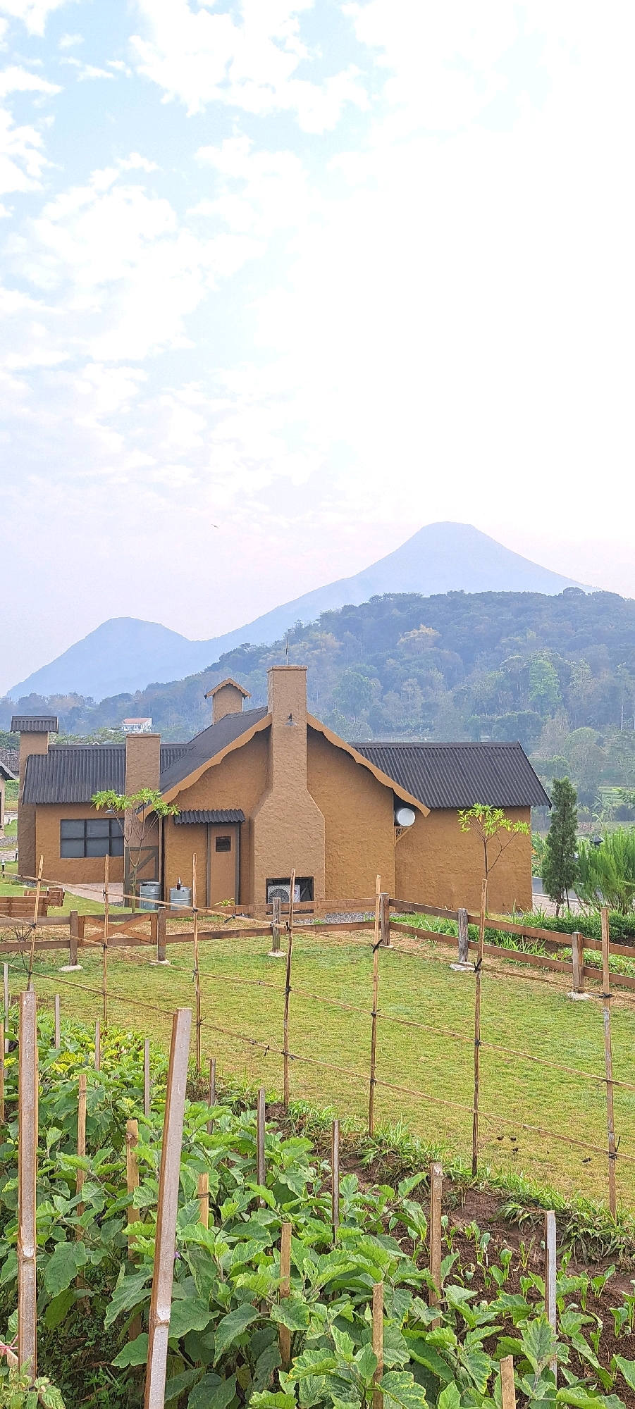 This is a vertical, eye-level, outdoor, daytime photograph of a rustic, tan-colored building with a dark, corrugated roof, set against a backdrop of lush green hills and distant mountains. In the foreground, a garden plot with rows of healthy green plants and bamboo stakes is visible, with a low wooden fence separating it from a grassy area. The building features a prominent brick chimney, several windows, and a satellite dish. A small tree grows near the building, and a single bird is in flight against the pale blue sky. The overall impression is one of rural tranquility, with the building appearing to be a house or a small resort. The location is identified as Tamiajeng, Indonesia.
