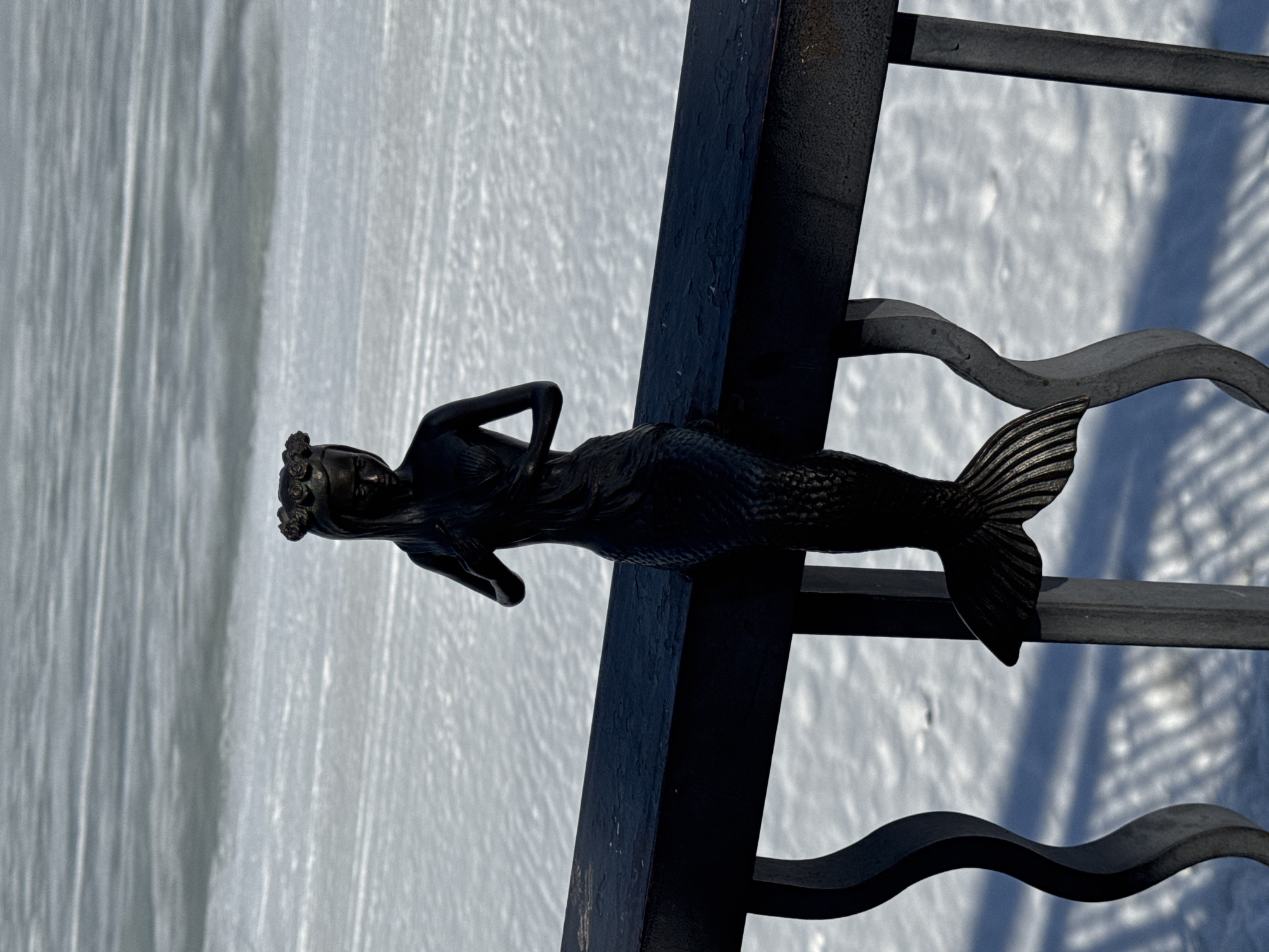 A dark metal mermaid statue is positioned on a metal structure in Chaikovsky, Russia. The statue features a human upper body with a fish tail and is adorned with a floral crown. The metal structure supporting the statue includes vertical and horizontal bars. The background is light and appears to be snowy or icy.
