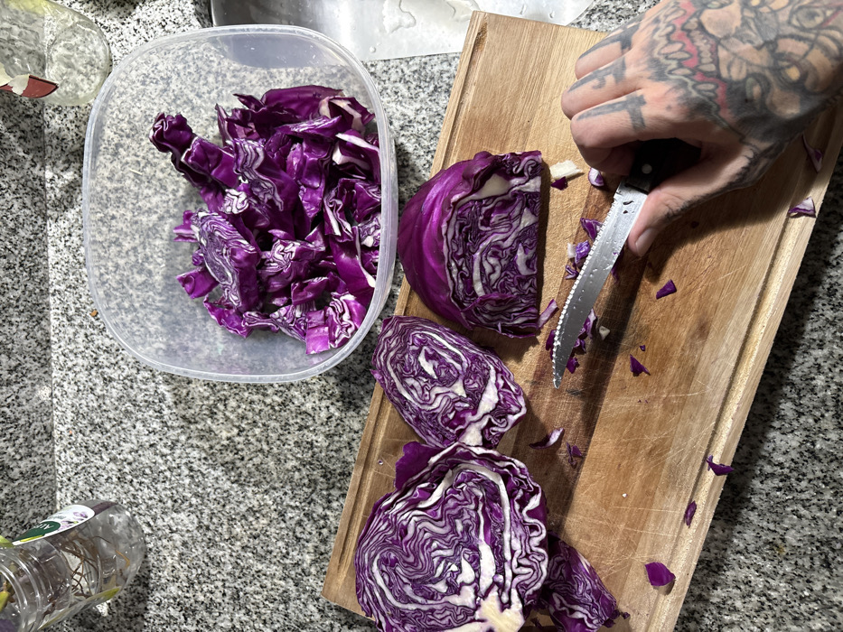 Chopped red cabbage fills a clear plastic container. On a wooden cutting board, more red cabbage is quartered, with loose pieces scattered around. A tattooed hand holds a serrated knife, positioned mid-cut. The scene is set on a gray speckled countertop, with a jar of herbs visible in the corner. Overhead lighting casts shadows on the wooden board, highlighting the red cabbage's vibrant color.