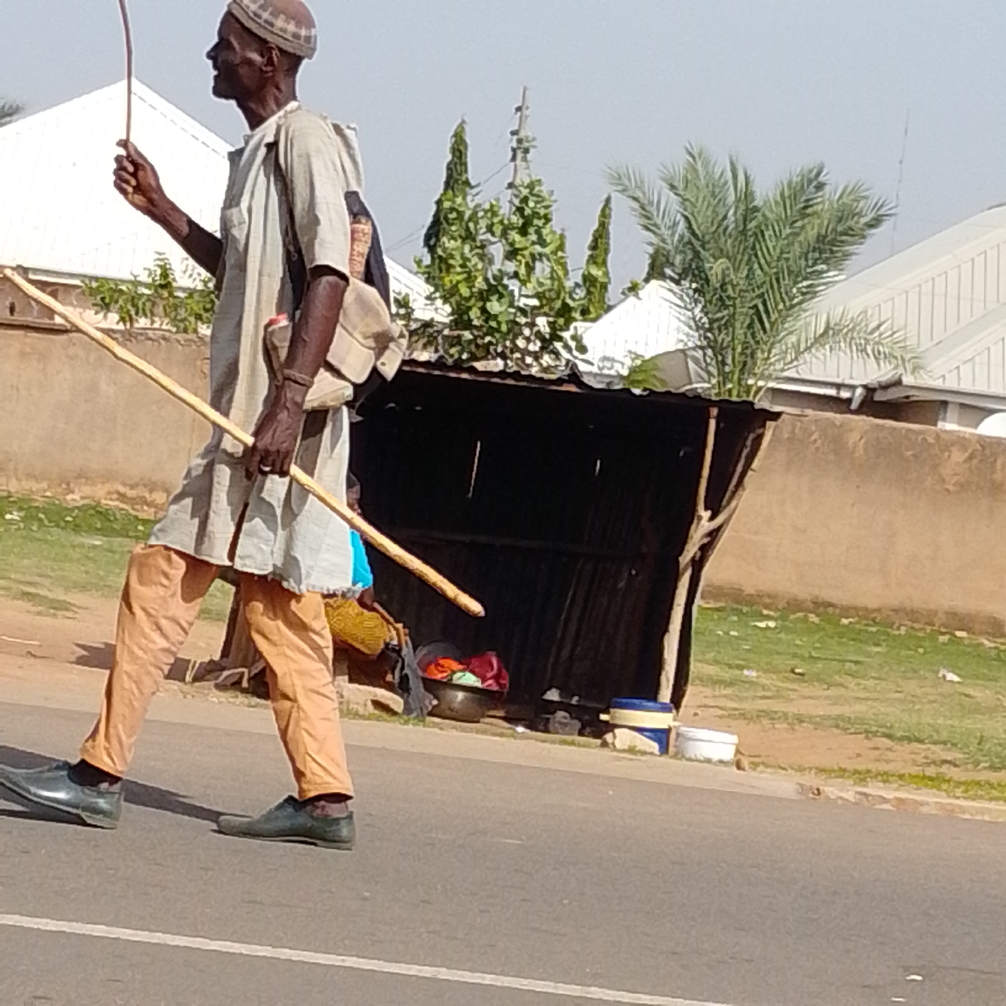 In Gombe, Nigeria, a lone elderly man walks on a paved road. He wears a traditional brown and white patterned cap, a light-colored loose-fitting tunic, and light brown trousers. He carries a long, thin wooden stick in his right hand and another, similar stick rests on his left shoulder, extending forward. A light brown satchel is slung over his left shoulder. The man is walking towards the left side of the frame. Behind him, a makeshift structure with a corrugated metal roof and dark walls stands. In front of this structure, various items are visible, including a metal bowl filled with colorful clothing and two cylindrical containers, one blue and yellow, the other white. The environment suggests an outdoor setting with a clear sky and scattered green vegetation.
