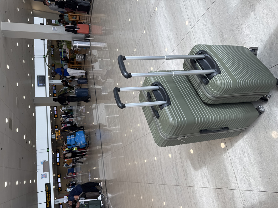 The image displays an active indoor airport terminal in Zanzibar, Tanzania. In the immediate foreground, two olive-green hardshell suitcases, a larger and a smaller one, exhibit horizontal ridged designs and extended silver telescoping handles with black grips. The larger suitcase features visible spinner wheels. The highly reflective light-toned tiled floor mirrors overhead lighting and the figures in the background.
The midground and background depict a busy environment with numerous individuals, consistent with airport travelers and staff. People are observed walking, standing, and engaging with luggage. Specific actions include a person pushing a blue luggage cart heavily laden with bags in the mid-left, and another individual leaning on a green luggage cart in the far left. A woman dressed in a black abaya walks in the central-right background. In the far right background, a lounge area with wooden tables and chairs contains seated individuals, positioned near a tall, cylindrical silver waste receptacle. Check-in counters or gates, distinctly labeled with numbers from 14 through 27, line the distant background, complemented by digital information display boards mounted on walls. The overarching environment features high white ceilings equipped with numerous recessed circular lights and large white structural pillars.