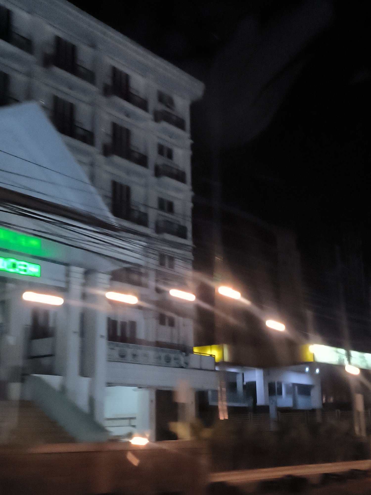 This blurry, motion-filled photograph captures an urban street scene at night, likely taken from a moving vehicle in Banda Aceh, Indonesia. The primary subject is a multi-story, light-colored building on the left, featuring several floors of dark, rectangular windows or balconies, suggesting a commercial or residential structure.
Numerous black overhead utility wires crisscross the mid-ground, running horizontally across the frame. Below these wires, a bright green glowing sign is partially visible, clearly displaying the letters "CE" and hinting at "ACE," possibly indicating "Banda Aceh."
The scene is illuminated by several elongated, bright yellow-orange light streaks, which are characteristic of streetlights or building lights captured with motion blur, emphasizing the movement of the camera. In the background and to the right, other blurred buildings, possibly commercial establishments, are also visible, marked by similar light streaks and faint signage. The sky is completely dark, confirming the nighttime setting. The overall effect conveys the experience of passing through an illuminated city street after dusk.