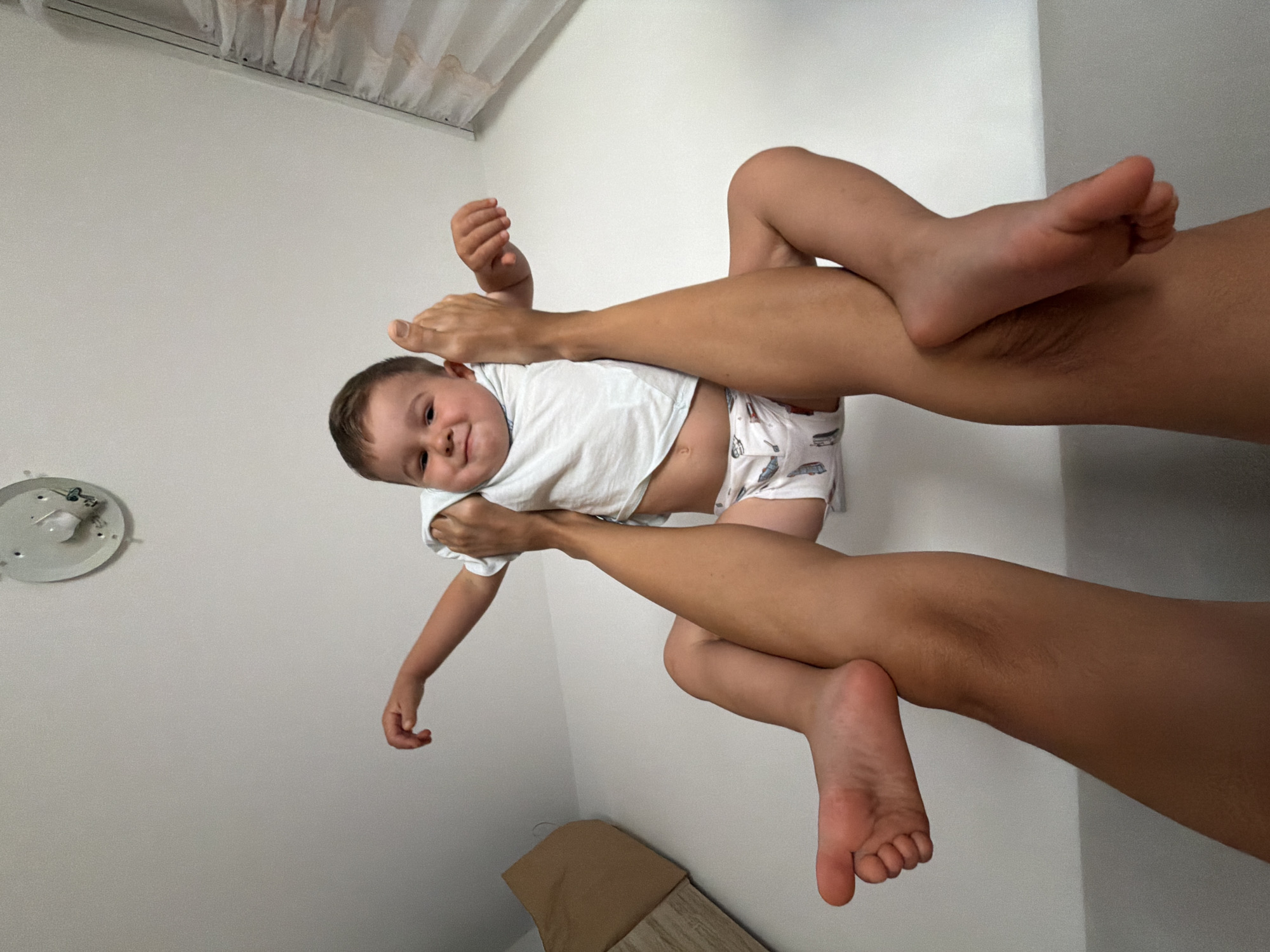 An indoor, vertically oriented shot captures a playful moment between an adult and a baby in Budva, Montenegro. The central subjects are a smiling baby and the legs and feet of an adult, positioned from a low angle looking upwards.
The baby, appearing to be a toddler with short light brown hair, is wearing a white short-sleeved t-shirt and white patterned shorts or a diaper cover. The baby is suspended in mid-air, held up by the adult's feet. The adult's bare legs and feet, tanned, are prominently featured, with one foot resting on the baby's chest area and the other foot seemingly supporting the baby's back or bottom, creating a cradle-like hold. The baby's arms are outstretched, and a wide, joyful smile is directed towards the viewer, indicating happiness and engagement in the playful activity.
The background consists primarily of plain white walls, suggesting a simple room interior. In the upper left corner, part of a white ceiling light fixture without a cover is visible. At the very top, a section of light-colored curtains or blinds can be seen. A piece of furniture, possibly a bed frame or bench with a brown cushion, is partially visible at the bottom right corner of the frame. The overall impression is a lighthearted and affectionate interaction between the adult and the child. No text is visible in the image.
