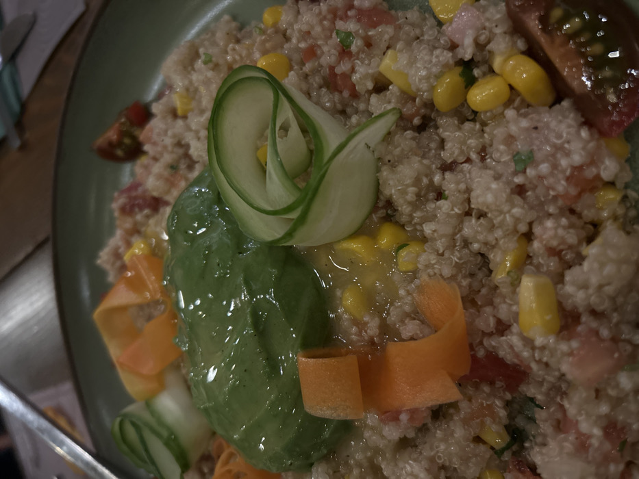 A close-up, top-down shot captures a vibrant quinoa salad presented in a green bowl. The main subject is the salad, composed primarily of fluffy white quinoa mixed with an assortment of colorful ingredients. Visible within the quinoa are bright yellow corn kernels, diced red tomatoes, and subtle green herbs, suggesting a fresh and healthy dish.
The salad is artfully garnished. A central feature is a thin, rolled slice of cucumber, shaped like a delicate rose. Adjacent to this, a large, smooth slice of avocado with a glossy, almost wet appearance, adds a creamy green element. Below the avocado and cucumber, bright orange carrot ribbons are visible, adding another pop of color and texture. The overall impression is one of careful preparation and presentation.
The lighting appears somewhat dim, suggesting an indoor setting, possibly a restaurant or a home, though no specific location details are visible in the image itself. A metallic utensil, possibly a fork or knife, is partially visible on the bottom left, hinting at a dining experience. There is no visible text in the image. The scene is static, showcasing the meal ready for consumption.