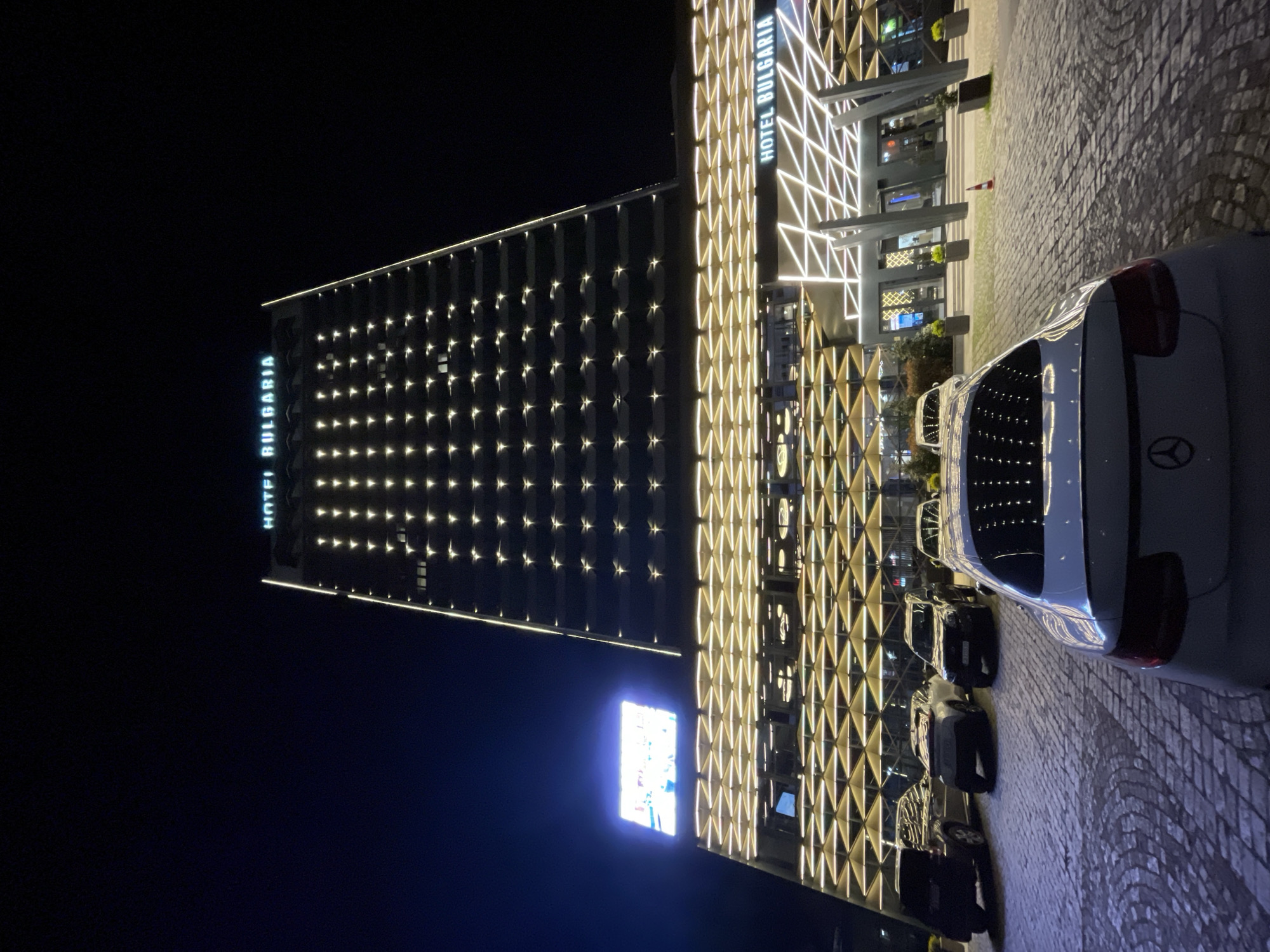 At night, the illuminated Hotel Bulgaria in Burgas, Bulgaria, stands tall against the dark sky. The building's façade features illuminated geometric patterns and vertical signage reading "HOTEL BULGARIA". The tower section of the hotel is punctuated with rows of evenly spaced lights. A line of parked vehicles is visible on the cobblestone pavement. A white Mercedes-Benz is partially visible on the right.