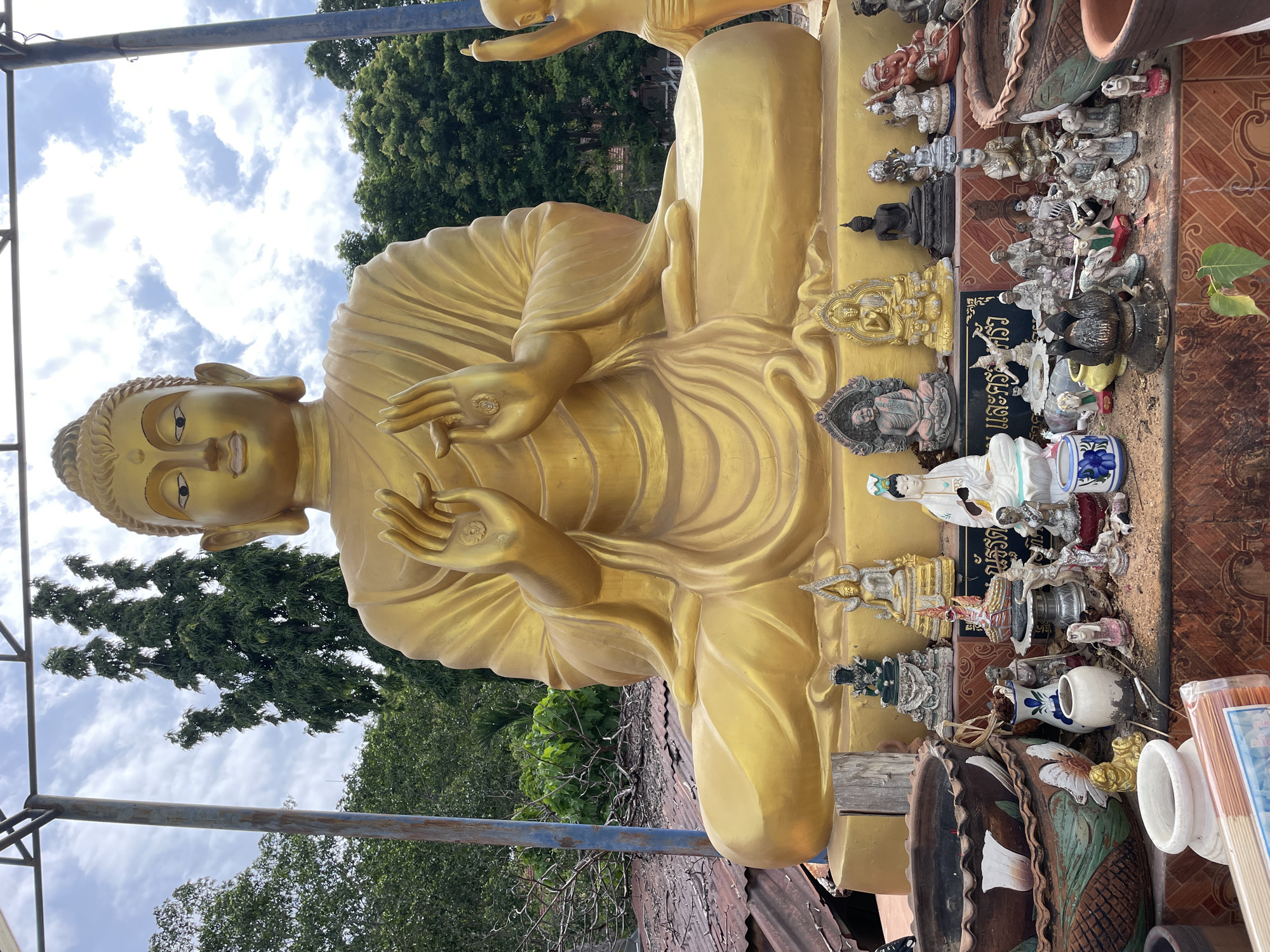 The image shows a large, golden Buddha statue sitting in a cross-legged pose. In front of the statue, there is a collection of smaller statues, offerings, and decorative items arranged on a tiered platform. These smaller items include figurines of Buddhas in various poses, other deities, animals, vases, and incense sticks. The scene is set outdoors, with trees and a cloudy sky visible in the background. Part of a metal structure, likely part of a shelter, is visible above the Buddha statue. The overall impression is one of a religious or spiritual site, possibly a temple or shrine, in Pattaya City, Thailand, given the inscription in Thai script on the platform.
