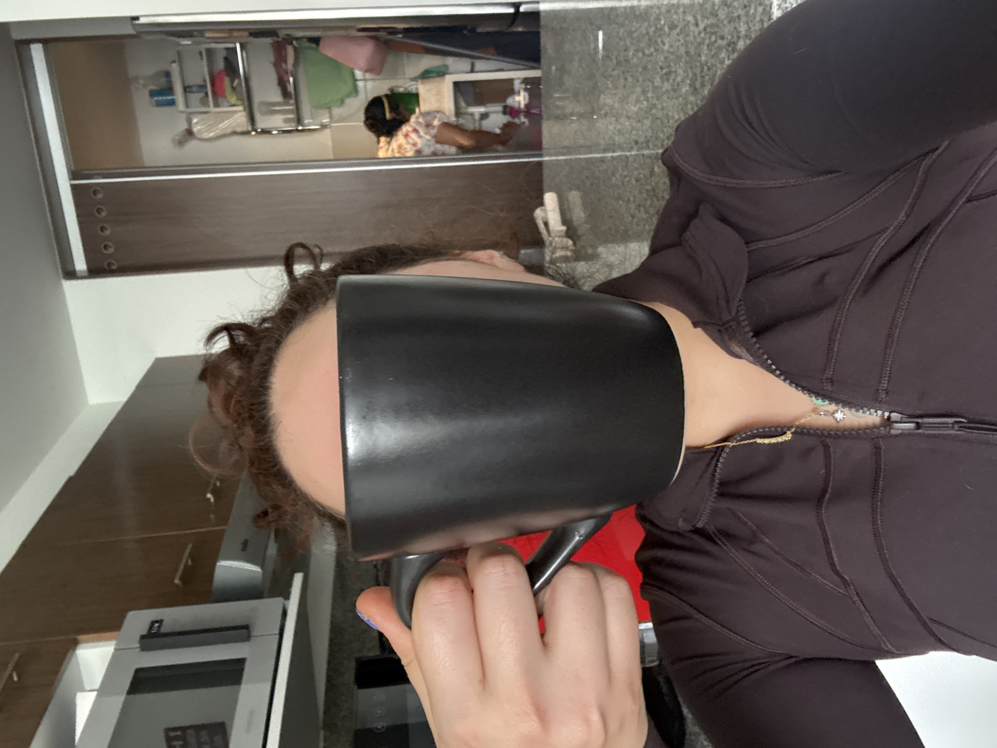 A woman is holding a black mug in front of her face. She's wearing a dark brown zippered jacket and is in a kitchen with brown cabinets and stainless steel appliances. A reflection in a mirrored cabinet shows a person in a floral shirt and apron. The location is Mexico City, Mexico.