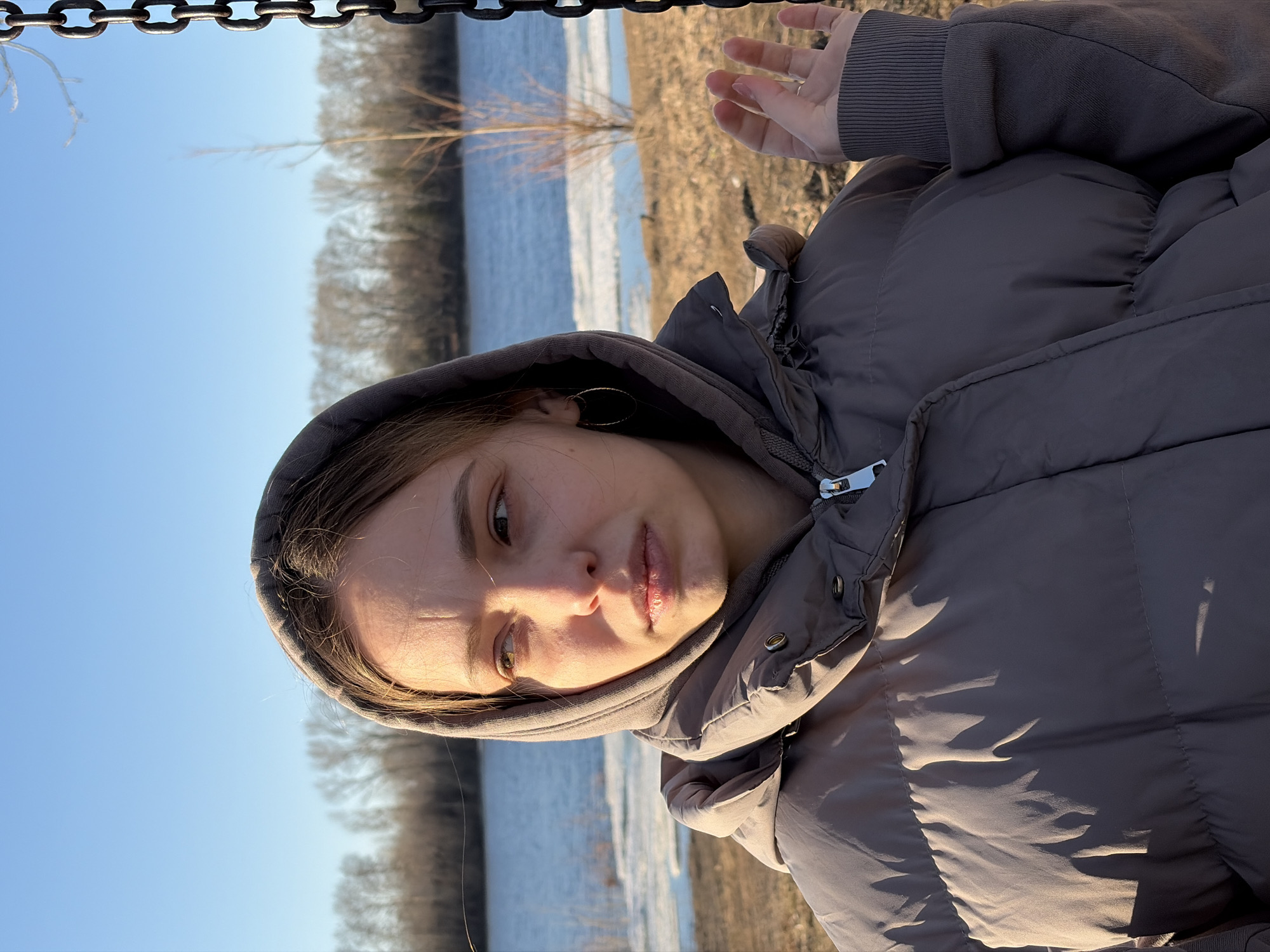 A woman wearing a gray hooded puffer jacket is positioned in the foreground of the image, with a body of water, possibly the Kama River, and shoreline visible in the background. The water appears to have patches of ice. Bare trees line the distant shore. A metal chain is visible at the top left of the frame. The sky is blue and clear. The woman is looking to her left and slightly downward. Part of her hand is in the upper right corner. The scene is likely taking place in Chaikovsky, Russia, given the river and typical landscape.