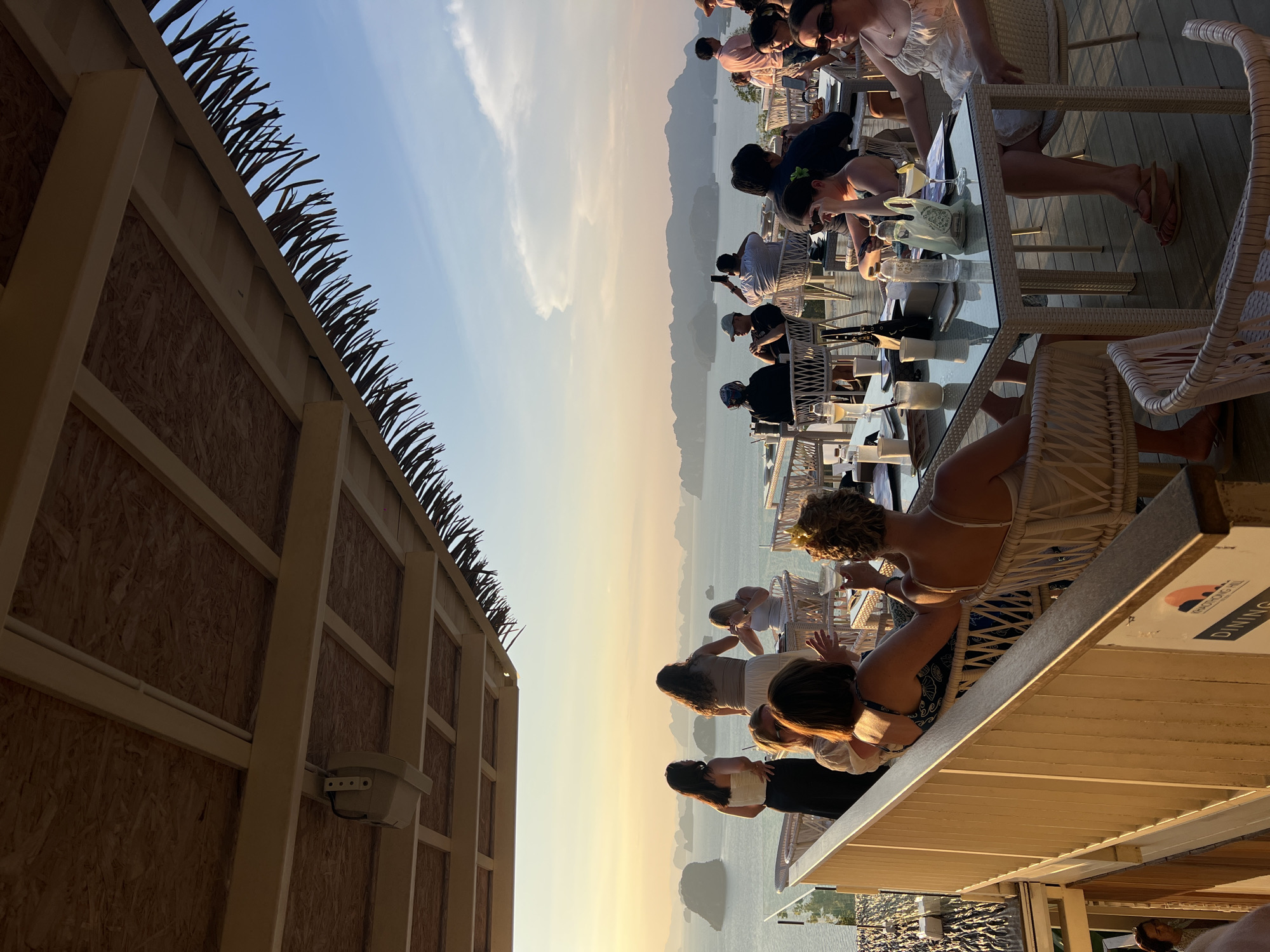 The image captures a scene in Khao Thong, Thailand, depicting an outdoor dining setting. A long row of patrons sit at tables with clear surfaces, likely enjoying a meal. In the background, the landscape includes visible islands and a hazy, golden-hued sky. Adjacent to the dining area is a building with a wooden facade and a roofline decorated with what appear to be thatched elements.