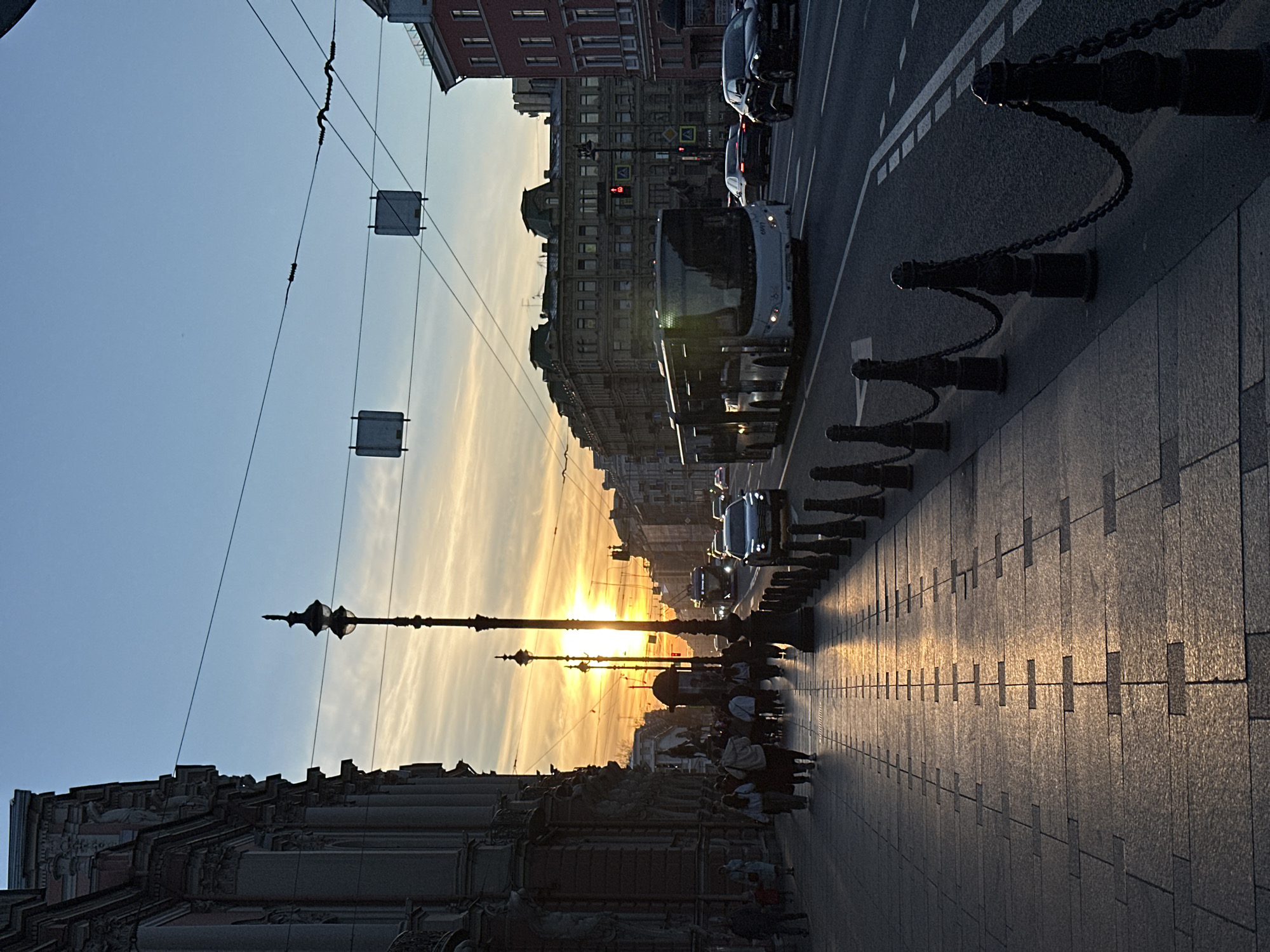 The image shows a street scene in Saint Petersburg, Russia, during sunset. The sun is visible in the distance, casting a golden glow along the Nevsky Prospekt, the main street. Several buses and cars are on the street. People walk along the sidewalk, which is paved with rectangular blocks. The sidewalk is bordered by a low wall of decorative black metal posts connected by chains. Electrical lines run above the street. The buildings on either side of the street are multistory and have detailed facades.