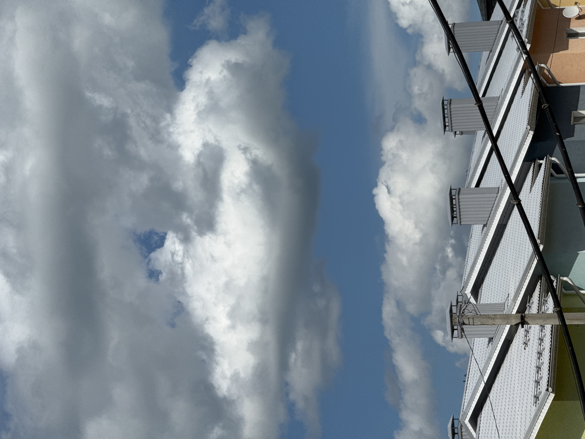 The image is a vertical, low-angle shot of a building against a cloudy blue sky. The building appears to be multi-story, with a light gray, textured roofline visible. Several rectangular, silver-colored ventilation units with angled tops are mounted on the roof, some appearing to be chimneys. Black electrical wires are strung across the frame, supported by a concrete pole with additional wiring. The building's exterior walls are painted in alternating bands of pale orange, dark gray, and pale green. Some windows are visible on the upper floors. The sky is filled with dramatic, voluminous white and gray clouds, suggesting a dynamic or possibly overcast weather condition. The lighting indicates it is daytime, likely mid-day or afternoon given the brightness. The scene is an outdoor urban or suburban setting, possibly residential or commercial. There is no visible text in the image.