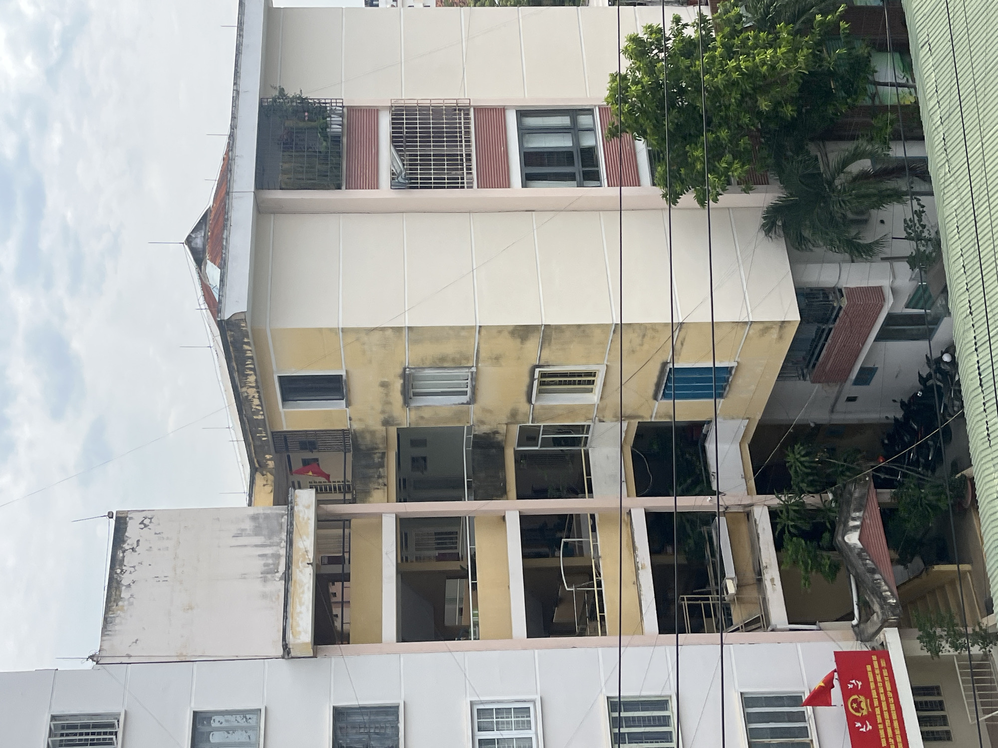 The image captures a multi-story building in Nha Trang Ward, Vietnam. The building's architecture is a mix of styles, with sections painted in white and yellow. The building shows signs of age and wear, with some areas exhibiting discoloration and staining. Windows are visible on all floors, some with security bars or screens. A Vietnamese flag is prominently displayed on one of the lower floors. Vegetation, including trees and potted plants, is interspersed around the building. Power lines crisscross the foreground. A neighboring building is partially visible on the right side of the frame. The sky is overcast.
