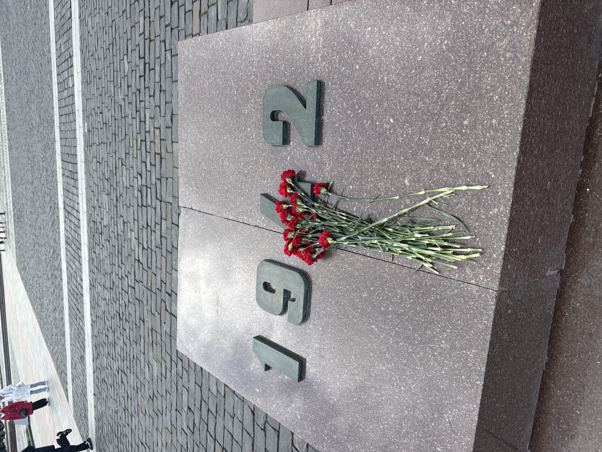 The image shows a close-up of a stone monument with the year "1942" inscribed on it in large, metallic letters. A small bouquet of red carnations has been placed on the monument. The monument appears to be situated on a paved area of gray brick. In the background, blurred figures of people and street objects are visible. Based on the monument's appearance and the year inscribed, this is likely a memorial related to World War II, specifically the Battle of Moscow in 1942. The location is Moscow, Russia.