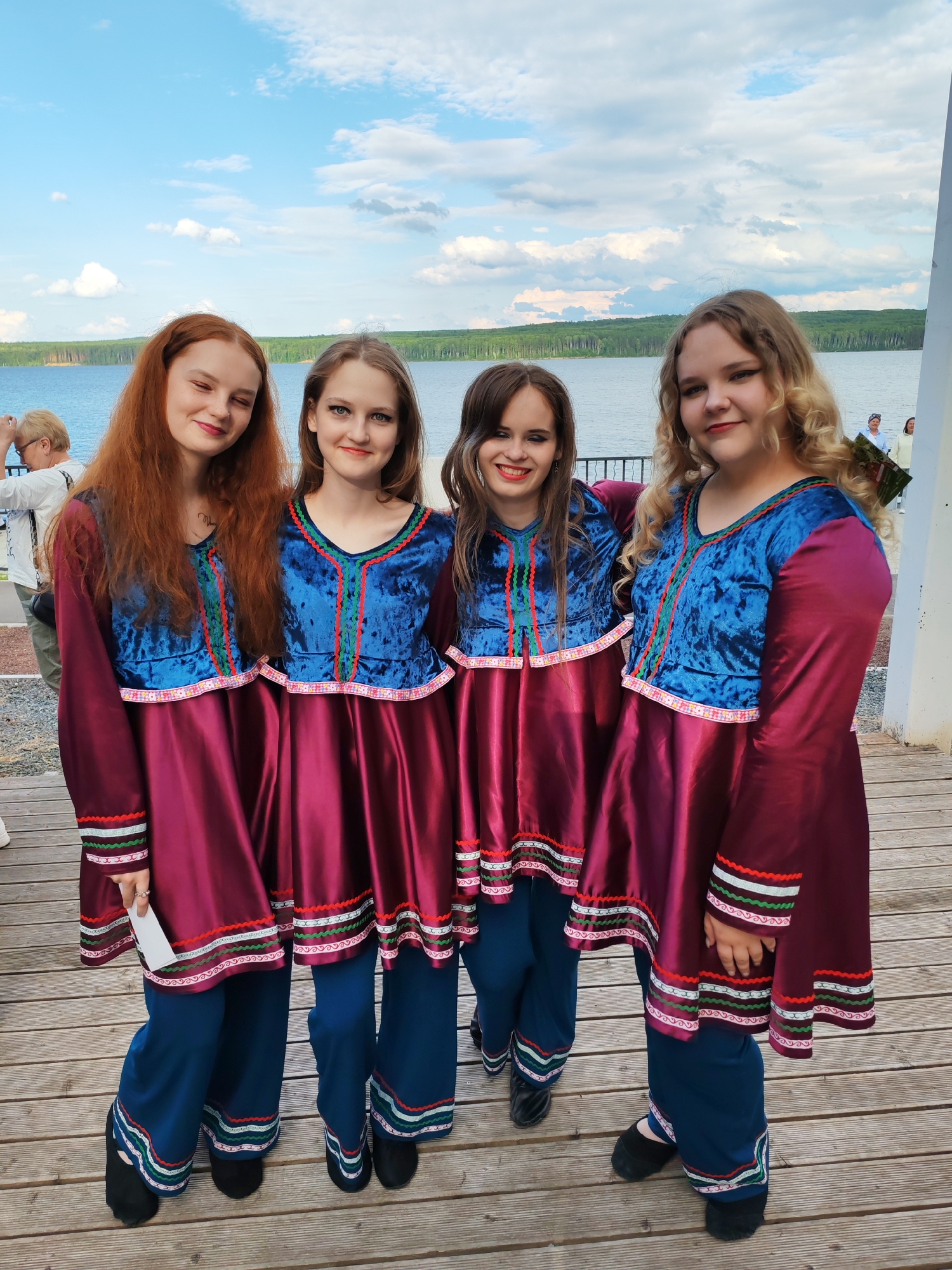 Four young women stand on a wooden deck, posing for a photograph. They are dressed in traditional outfits consisting of a dark red tunic with blue velvet accents on the chest and sleeves, adorned with colorful striped trim. They also wear matching blue pants. The women have varying hair colors, ranging from red to blonde, and are smiling at the camera.
Behind them, a body of water, likely a lake, stretches to the horizon, with a line of trees in the distance. The sky is filled with fluffy white clouds against a clear blue backdrop, suggesting a pleasant day. The presence of other people in the background and the clear sky indicate it is likely daytime. The setting appears to be an outdoor gathering or festival by the water. One woman on the far left is holding a white piece of paper or card. The overall mood is cheerful and festive.