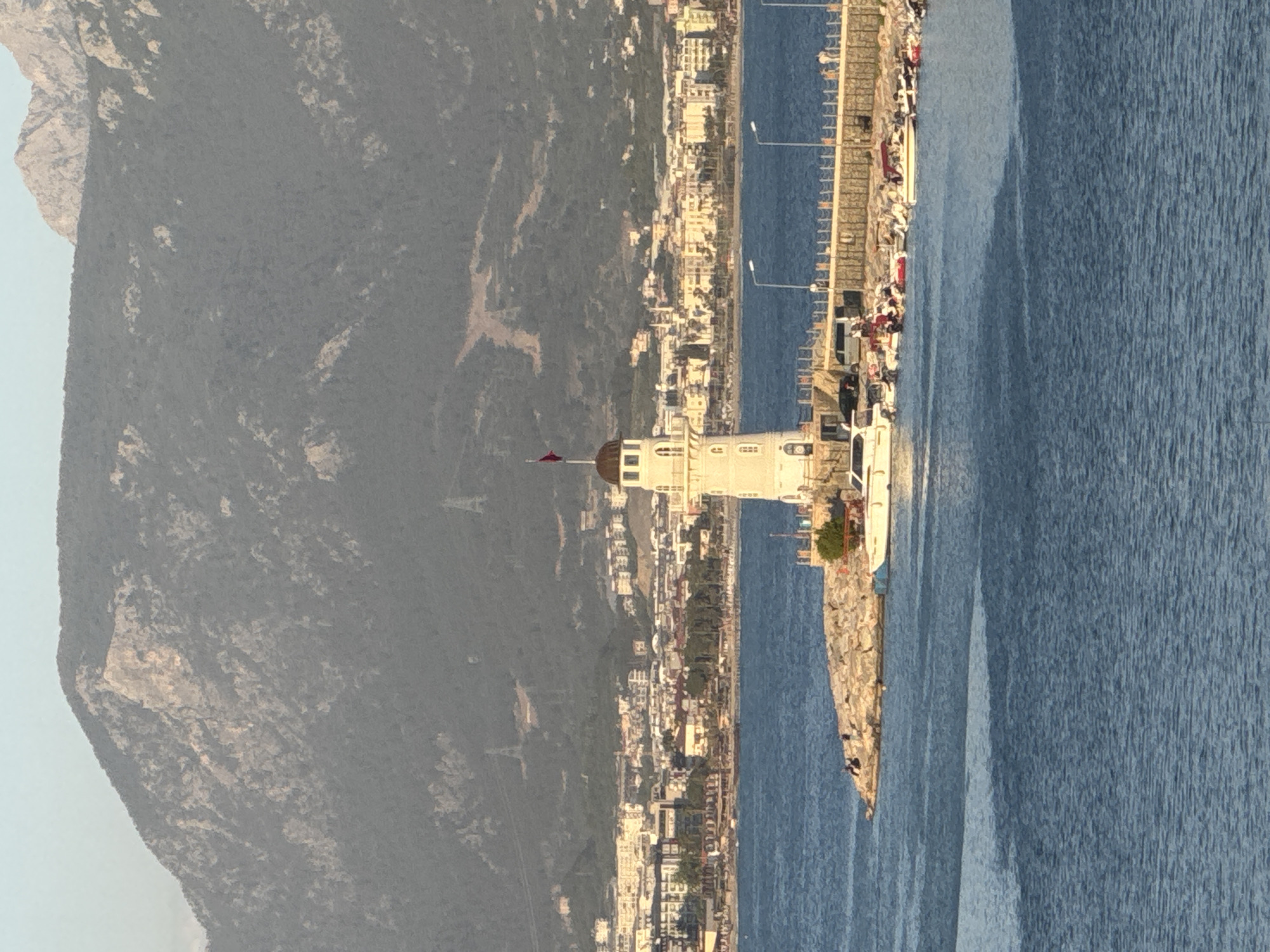 The image depicts a coastal scene in Alanya, Turkey. In the foreground, the Alanya lighthouse sits on a rocky breakwater extending into the blue water of the Mediterranean Sea. A red Turkish flag is visible atop the lighthouse. Along the breakwater, people are walking and fishing. A pier is visible with boats docked alongside, and street lamps illuminate the area. Behind the lighthouse and breakwater is the city of Alanya, with buildings extending along the coastline. The backdrop features a large mountain with sparse vegetation. The sky is clear and suggests daytime.