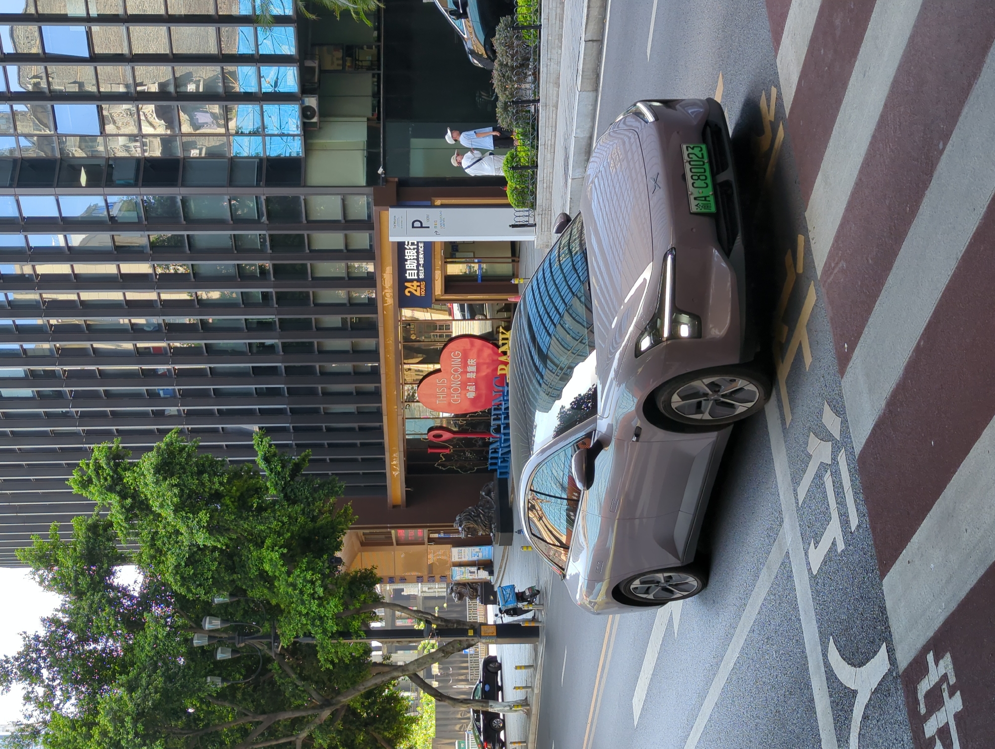 The image shows a gray electric sedan with license plate A-C80023 driving on a street in Yuzhong District, China. The car is in the foreground, facing the viewer and positioned on a crosswalk. The street is paved and has yellow and white lane markings. In the background, there is a modern glass-fronted building with vertical dark framing. To the left of the building, there is a street lamp with multiple lights and lush green trees. A scooter with a blue box on the back is parked near the sidewalk. There are two people walking on the sidewalk in front of the building. The building's facade reflects the surrounding environment, including other buildings and trees. A red circular sign with white text is visible on the building.