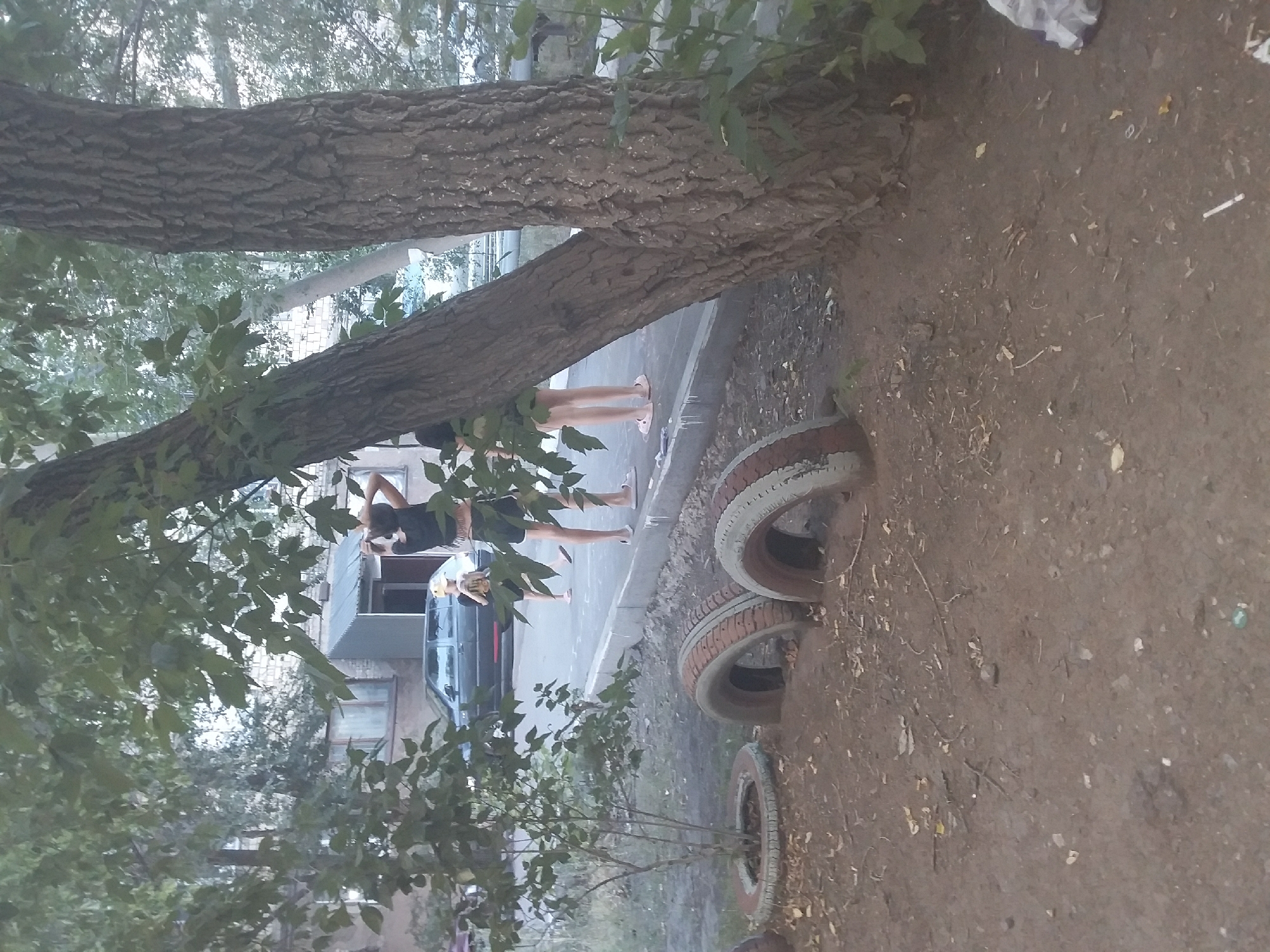 The image depicts a street scene in the city of Saran, Kazakhstan. In the foreground, several old tires are partially buried in the dirt, some containing plants. Two large trees dominate the right and left sides of the frame, with foliage obscuring parts of the background. Behind the tires, a paved walkway runs along the edge of the street. A dark-colored car is parked on the street. Three individuals are visible on the walkway. One person, dressed in black shorts and a black top, stands with their back to the camera, hands raised near their head. To their left, two younger individuals, one wearing a yellow cap, are also present. Buildings with visible windows are in the background. The ground is covered with dirt, dry leaves, and scattered debris.