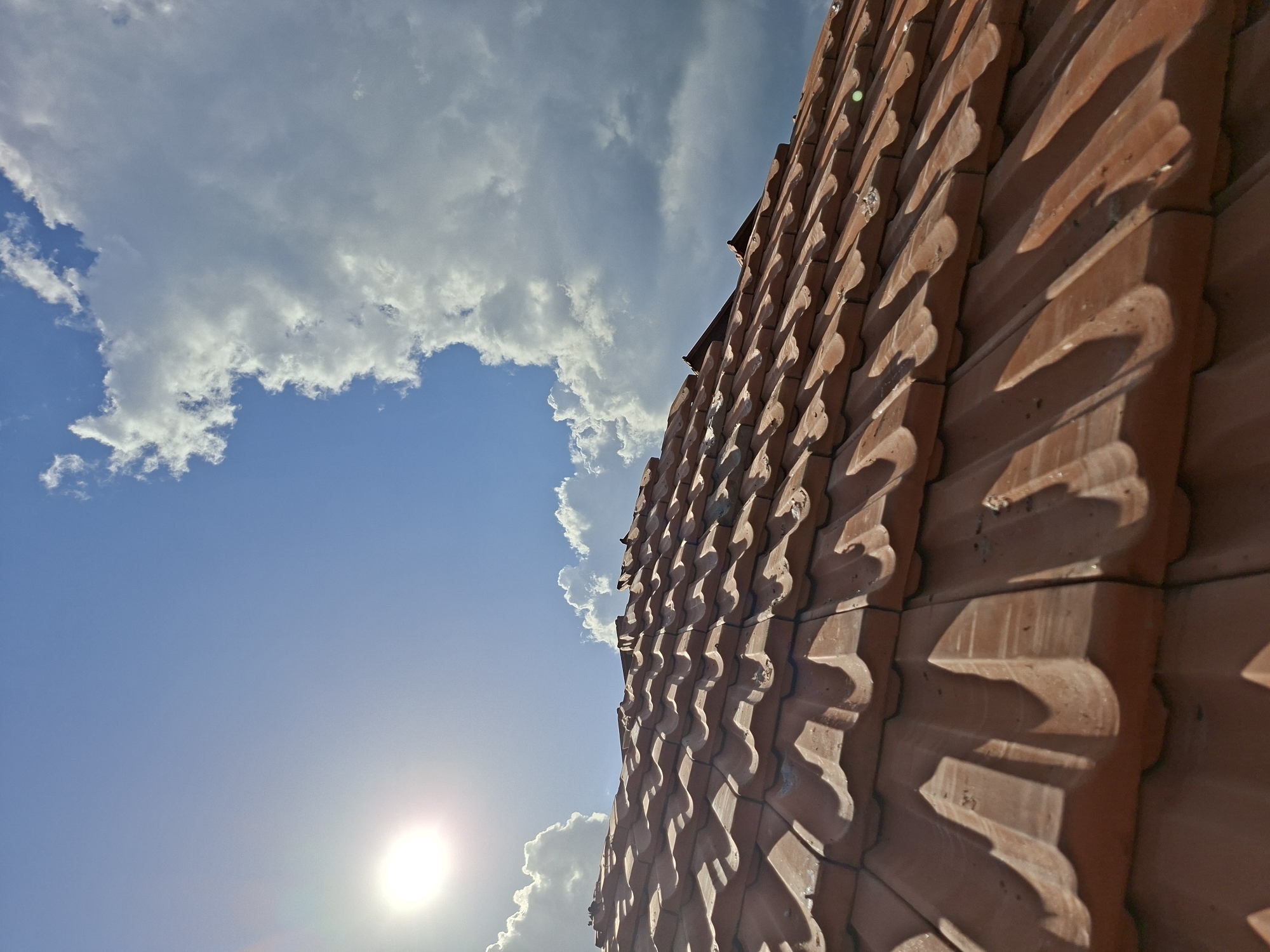 The image shows a close-up, low-angle view of a roof with terracotta tiles against a blue sky with scattered white clouds. The sun is visible, casting a glare and highlighting the texture of the tiles. The location is Bojuritsa, Bulgaria. There are no people visible. The primary subject is the roof tiling.