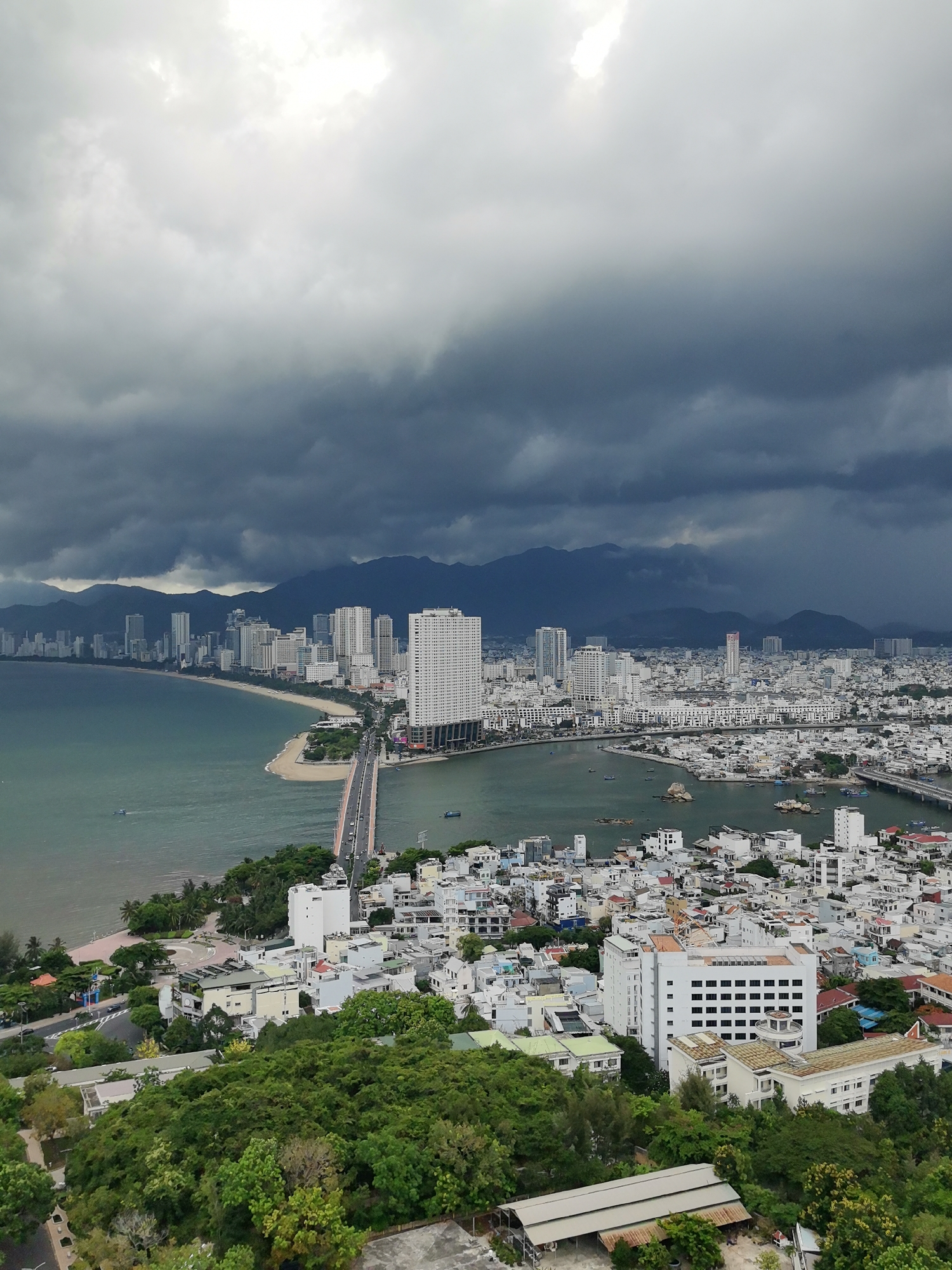 This aerial, high-angle shot captures a coastal city in Vietnam, likely Nha Trang, under a dramatic, cloudy sky. The scene is dominated by a wide expanse of the ocean on the left, transitioning to a sandy beach and then a dense urban landscape. Numerous tall, white buildings, indicative of a city, line the coastline, with a prominent bridge connecting different parts of the city over a body of water. Further inland, a range of mountains forms the backdrop, shrouded in the dark, stormy clouds that suggest an impending storm or recent rain.
The city appears to be a bustling coastal hub with a mix of modern high-rises and lower-rise buildings. Lush green trees and vegetation cover the foreground hill, contrasting with the predominantly white and grey tones of the city. Several small boats are visible on the water, suggesting ongoing activity despite the weather. The overall mood is somewhat somber due to the dark clouds, yet the vibrant green of the foliage and the expanse of the sea offer a sense of natural beauty. The time of day is indeterminate but leans towards overcast daylight. There is no visible text in the image.