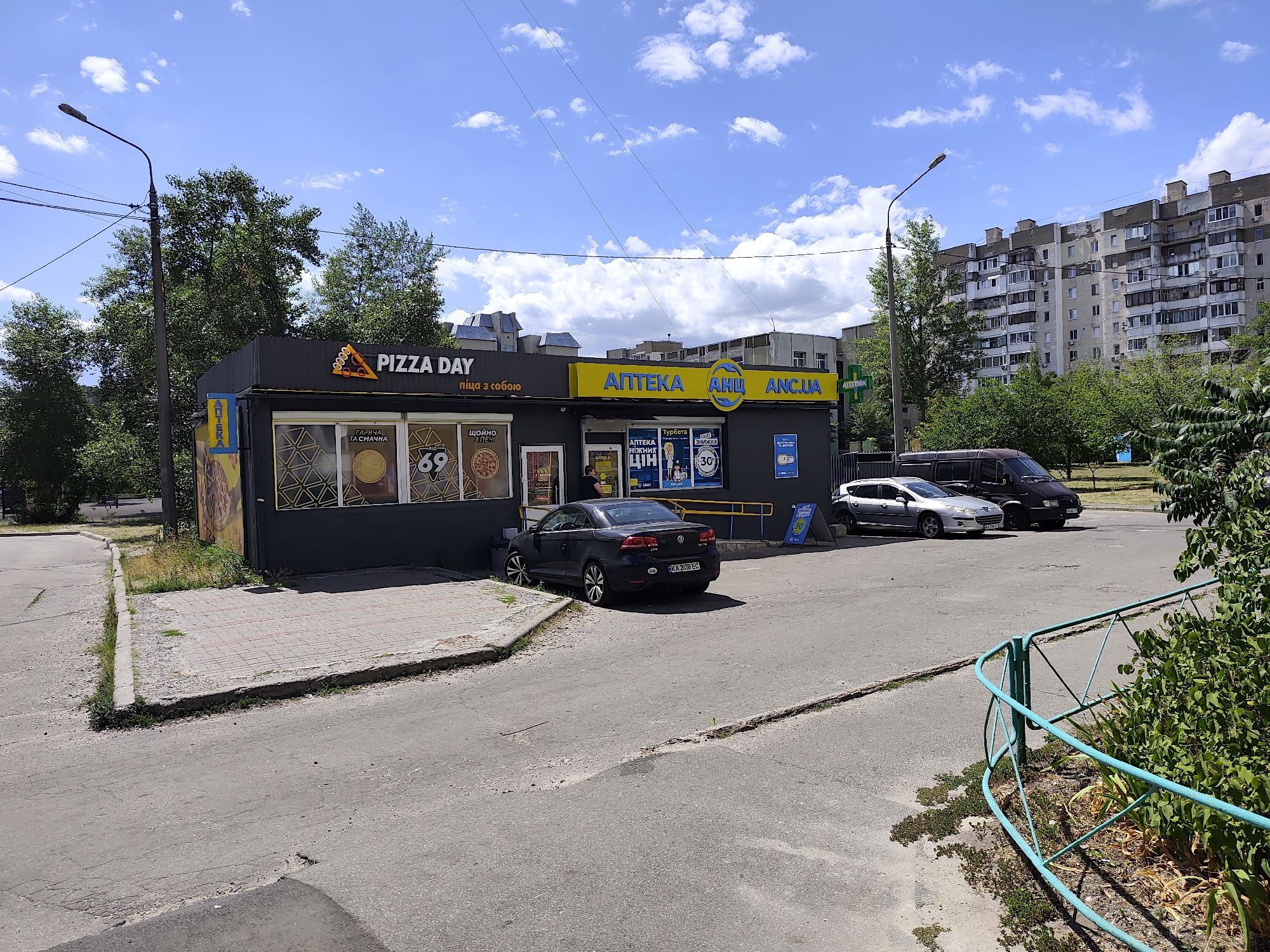 The image shows a street scene in an urban environment under a blue sky with scattered clouds. In the foreground, there is a paved road with some cracks and a section of discolored asphalt. To the right, a turquoise metal fence encloses a patch of green vegetation.
Dominating the middle ground is a small commercial building with dark gray siding and a prominent yellow awning. The building houses two businesses: "PIZZA DAY" on the left, indicated by a large sign with a stylized pizza slice, and "АПТЕКА АНЦ ANC.UA" on the right, a pharmacy with a yellow sign. Both businesses have display windows showcasing advertisements and products. A black Volkswagen sedan with a license plate reading "KA 2038 EC" is parked in front of the building, partially blocking the sidewalk. Adjacent to the sedan, a silver hatchback is parked, followed by a dark-colored van.
In the background, a multi-story residential building with numerous balconies and windows stands tall. Trees with green foliage are scattered throughout the scene, providing shade. A street lamp is visible on the left side of the image, and power lines are strung overhead. The overall impression is that of a typical neighborhood commercial area.