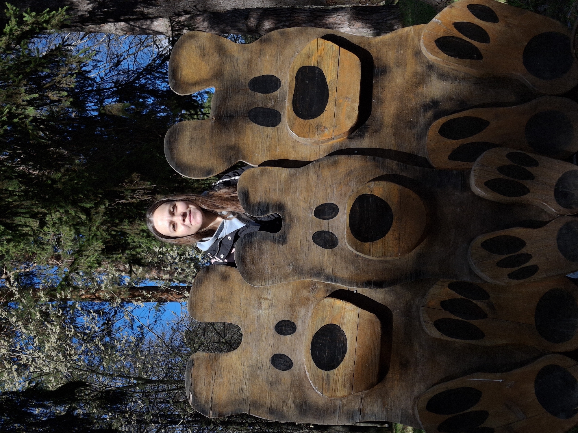 A woman poses in front of a vertical wooden structure featuring three stacked bear figures in a park in Sarapul, Russia. The figures are stylized with large round heads and paw prints, made of light brown wood with dark brown painted features for eyes, noses, and paw pads. The woman is light-skinned with long brown hair, and she's wearing a black and light blue jacket. She stands behind the middle bear, her face visible through the cutout. The background shows trees and a blue sky. The lighting is bright.