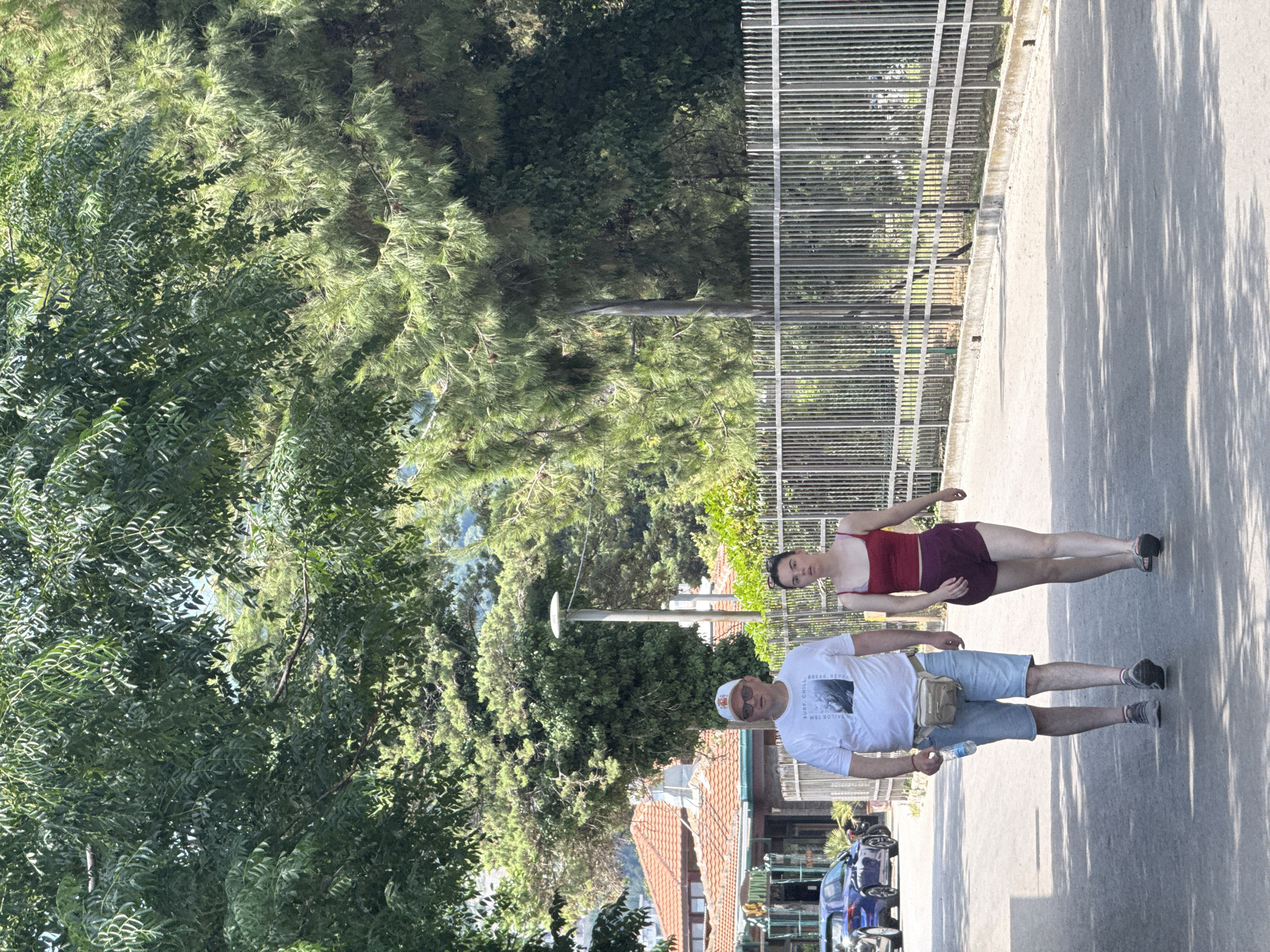 Two individuals, a man and a younger woman, walk side-by-side on a paved path, possibly in the city of Boreti, Montenegro, given the context. The man, wearing a white t-shirt, denim shorts, a white baseball cap, and sunglasses, carries a light brown fanny pack around his waist and a water bottle in his right hand. The woman, dressed in a red tank top and maroon shorts, walks with her hands by her sides. They are on a paved pathway bordered by a tall, silver metal fence on the right. Behind the fence, dense green foliage and trees create a natural backdrop. To the left of the path, a building with a red-tiled roof and a blue car are visible, suggesting a semi-urban or resort setting. The lighting indicates it is daytime with dappled sunlight filtering through overhead trees, casting shadows on the path.