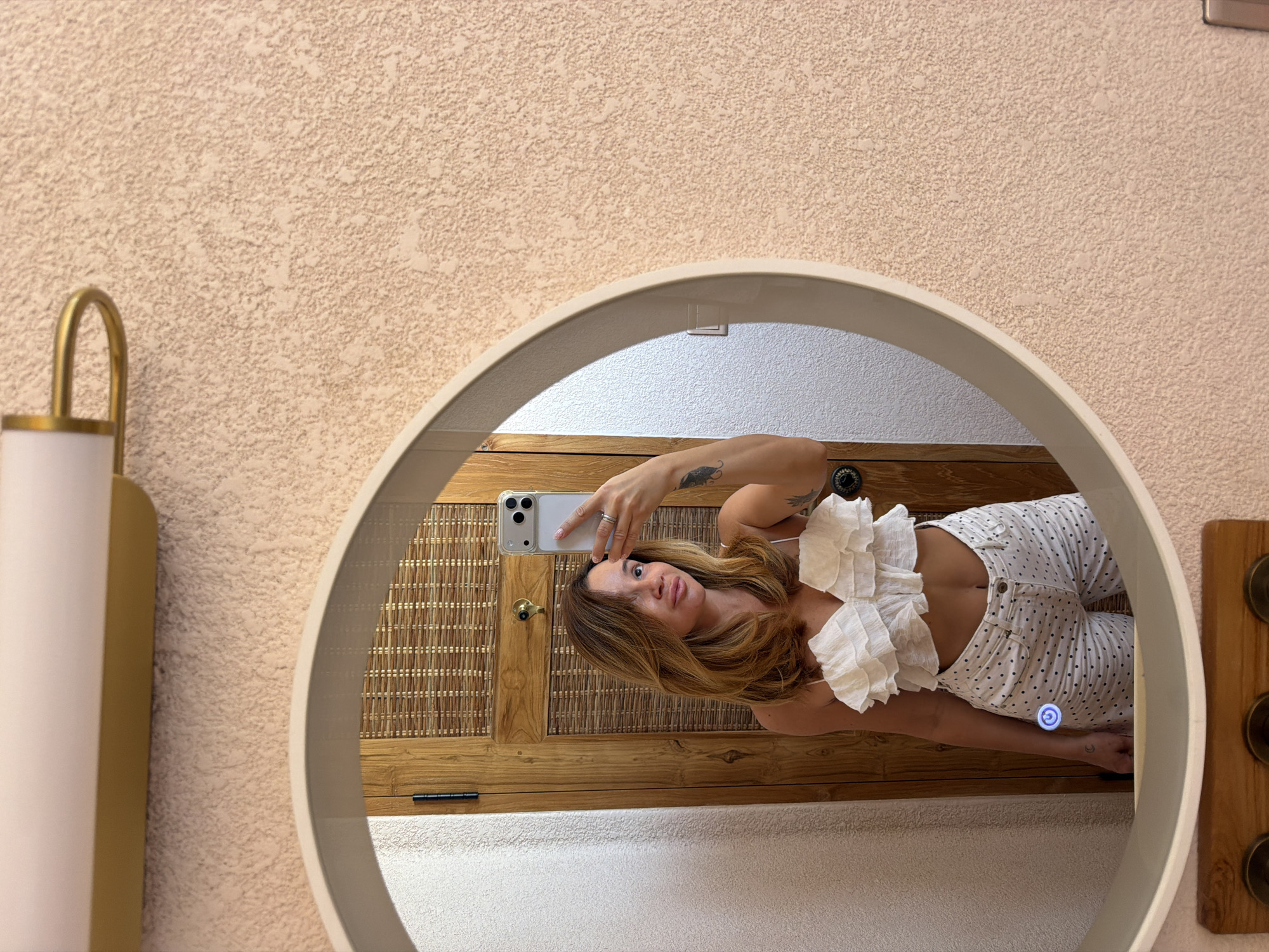 A young woman is taking a selfie in a round mirror. She is wearing a white ruffled top and white polka dot pants. She has long, wavy brown hair and is wearing rings on her fingers. The mirror reflects a wooden door with a woven insert and a light fixture above it. The wall behind the mirror is textured and pinkish-beige. The overall scene appears to be in a bathroom.