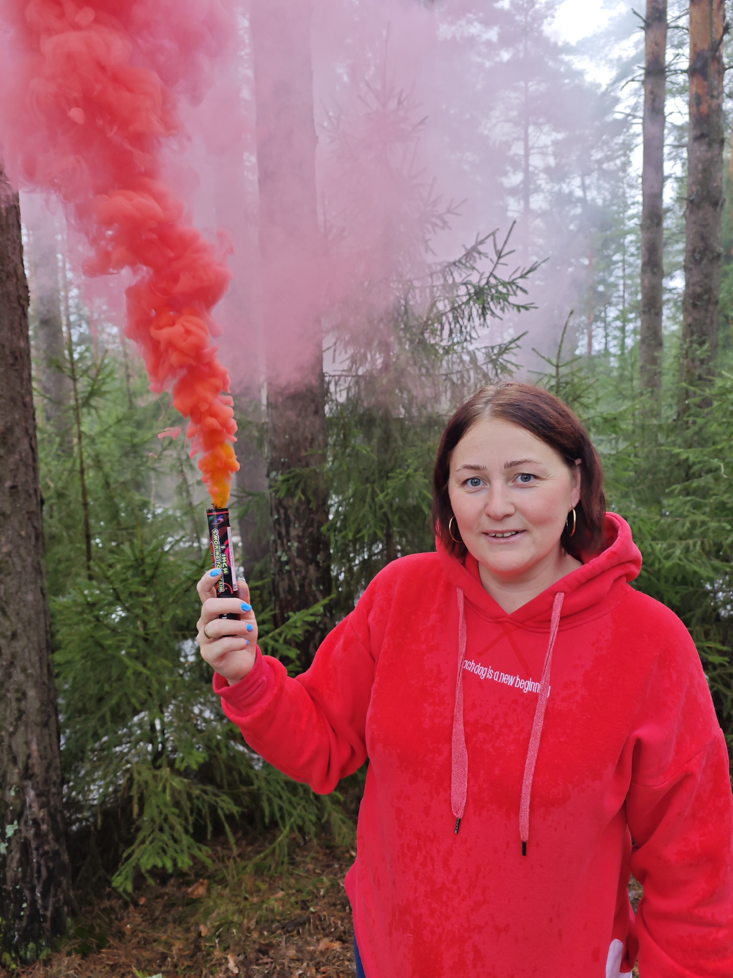 A woman stands in a forest near Markovsky, Russia, holding a lit red smoke grenade. She wears a red hoodie, gold hoop earrings, and has blue nail polish. The forest environment consists of coniferous trees and a leaf-covered ground. The sky appears overcast.