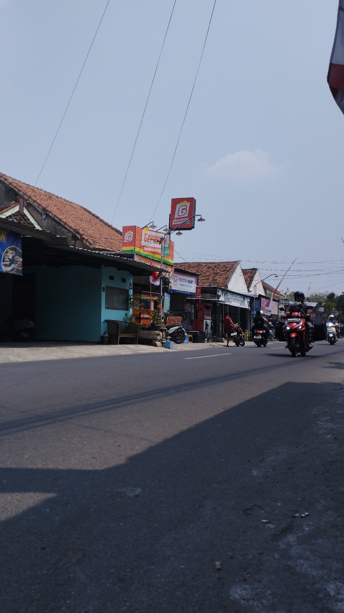 The image depicts a street scene in Ronowijayan, Indonesia, on a bright, clear day. The street is paved and appears to be a main thoroughfare, with several buildings lining the right side. These buildings are low-rise structures with tiled roofs, suggesting a commercial area. Signs advertising businesses like "GEPREK" and "MADURA MART" are visible, indicating retail establishments. Several motorcycles are present, with some being ridden and others parked. Two people are walking on the sidewalk in front of the businesses, and another person is on a motorcycle in the foreground. Power lines are strung across the sky above the buildings. The overall impression is of a bustling, everyday street in a Southeast Asian town.