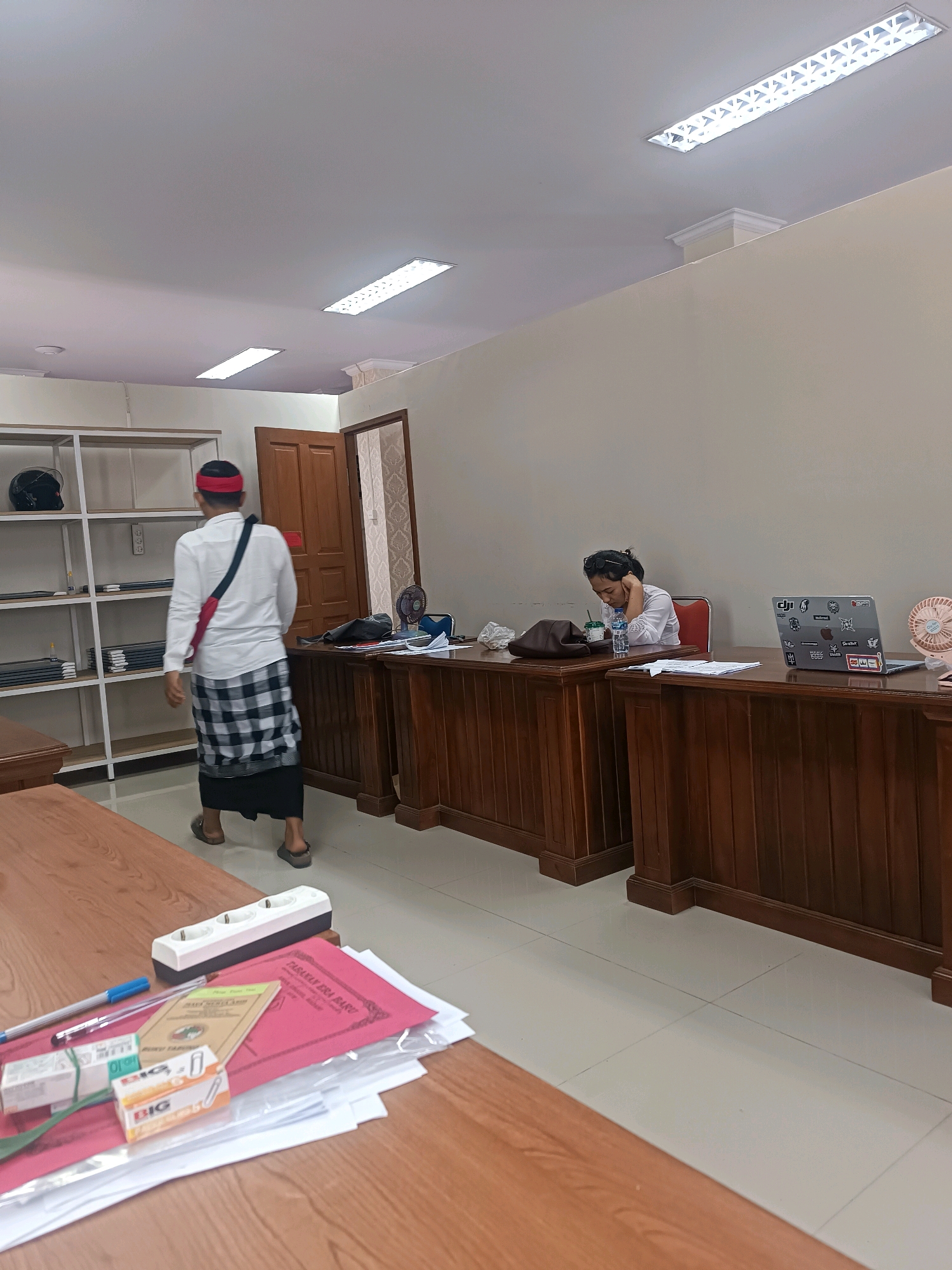 The image depicts an interior scene, likely an office or meeting room, within the city of Tabanan, Indonesia. The room is furnished with wooden desks, shelving units, and a tiled floor. The lighting fixtures on the ceiling are modern and fluorescent.
In the foreground, a wooden table is laden with various office supplies, including pens, papers, and a power strip.
To the left, a person wearing a white long-sleeved shirt, a red headband, and a black and white checkered sarong walks away from the camera, carrying a bag over their shoulder. A black helmet is visible on a shelf unit behind them.
To the right, a woman is seated at a desk, leaning her head on her hand, appearing to be tired or contemplative. A laptop with stickers and a small pink fan are on the desk in front of her. A bottle of water and a purse are also present.
The overall atmosphere suggests a working environment, possibly during a break or a period of quiet activity. The presence of a sarong suggests a cultural context specific to the region.