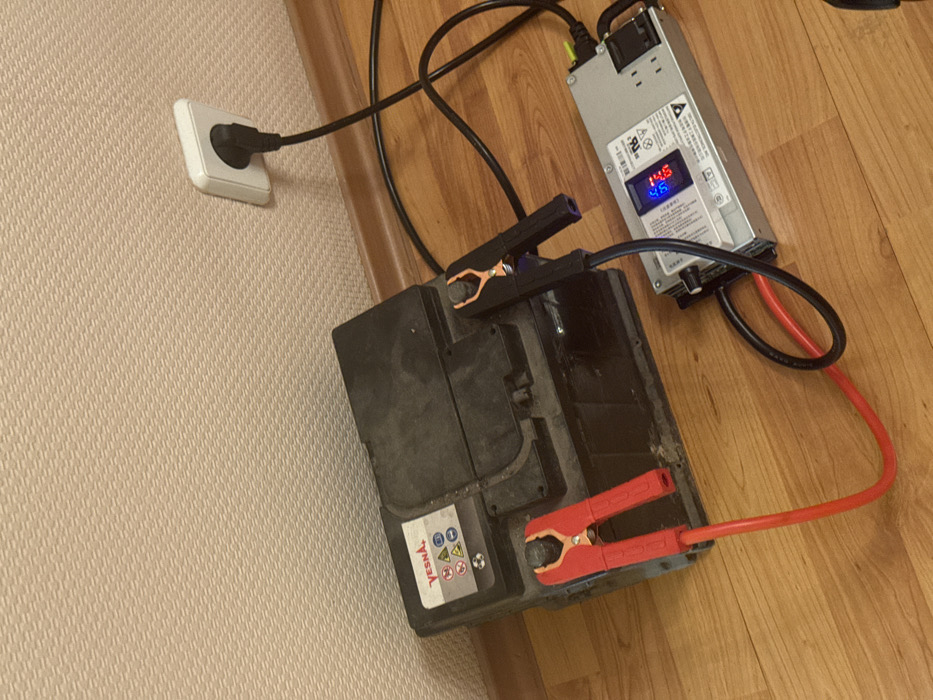 The image shows a black car battery on a wooden floor. Red and black jumper cables are connected to the battery terminals with alligator clips. The cables lead to a silver power supply with a digital display showing "14.6" and "4.6". The power supply is plugged into a white electrical outlet on a textured wall.