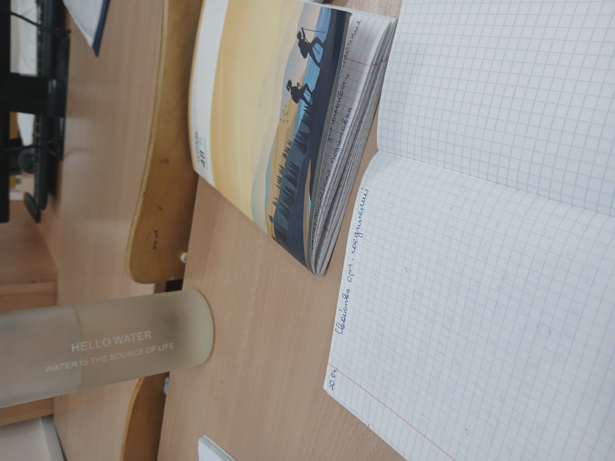 The image shows a desk at an angle. On the desk is a light brown water bottle with the words "HELLO WATER" and "WATER IS THE SOURCE OF LIFE" printed on it. Next to it, a notebook with a graphic of two hikers in a landscape is partially visible. An open notebook with squared paper lies open next to the notebook with the graphic. There is handwriting on the squared paper in Russian, including the date "20.04" and the phrase "Свойства орг. соединений". There is a keyboard and desk chair to the left of the water bottle.
The location is not discernible from the image. The notebooks are in Russian, which would suggest the notebooks are in a Russian-speaking area but does not indicate the city in which the image was taken.