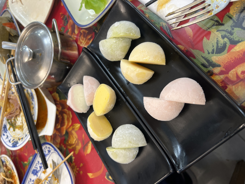 Two black plates display colorful mochi, each cut in half. Flavors include light green, yellow, and pink. A small metal pot with a lid sits near one plate. Chopsticks rest across the scene. A fork lies near the upper plate, indicating that the mochi is ready to be enjoyed. The surrounding surfaces are cluttered with other plates and bowls, suggesting a shared meal. The table is covered with a floral tablecloth.