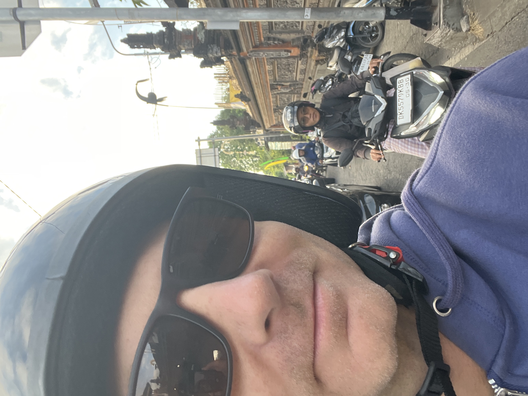 In Abiansemal, Indonesia, a man wearing a black helmet and sunglasses takes a selfie from the perspective of riding a scooter, his left shoulder and chest visible in the foreground wearing a purple hoodie. Behind him, a woman in a helmet and glasses rides another scooter, wearing a dark jacket over a checkered sarong. Several other scooters and people are visible on a street lined with traditional Balinese architecture. The scene is illuminated by natural light, suggesting it is daytime.