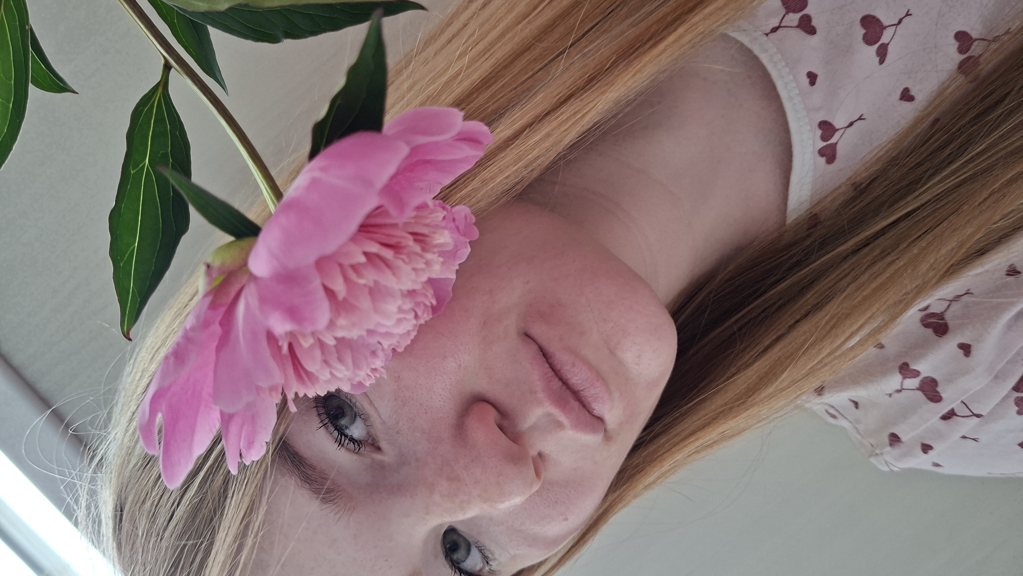 A close-up shot focuses on a young Caucasian woman's face, partially obscured by a large pink peony flower and its green leaves. The woman has fair skin with freckles, light blonde hair, and green eyes. She is wearing a light-colored shirt with a pattern of red hearts. The background is a neutral, light-colored wall. There is no discernible location in Chaikovsky, Russia.