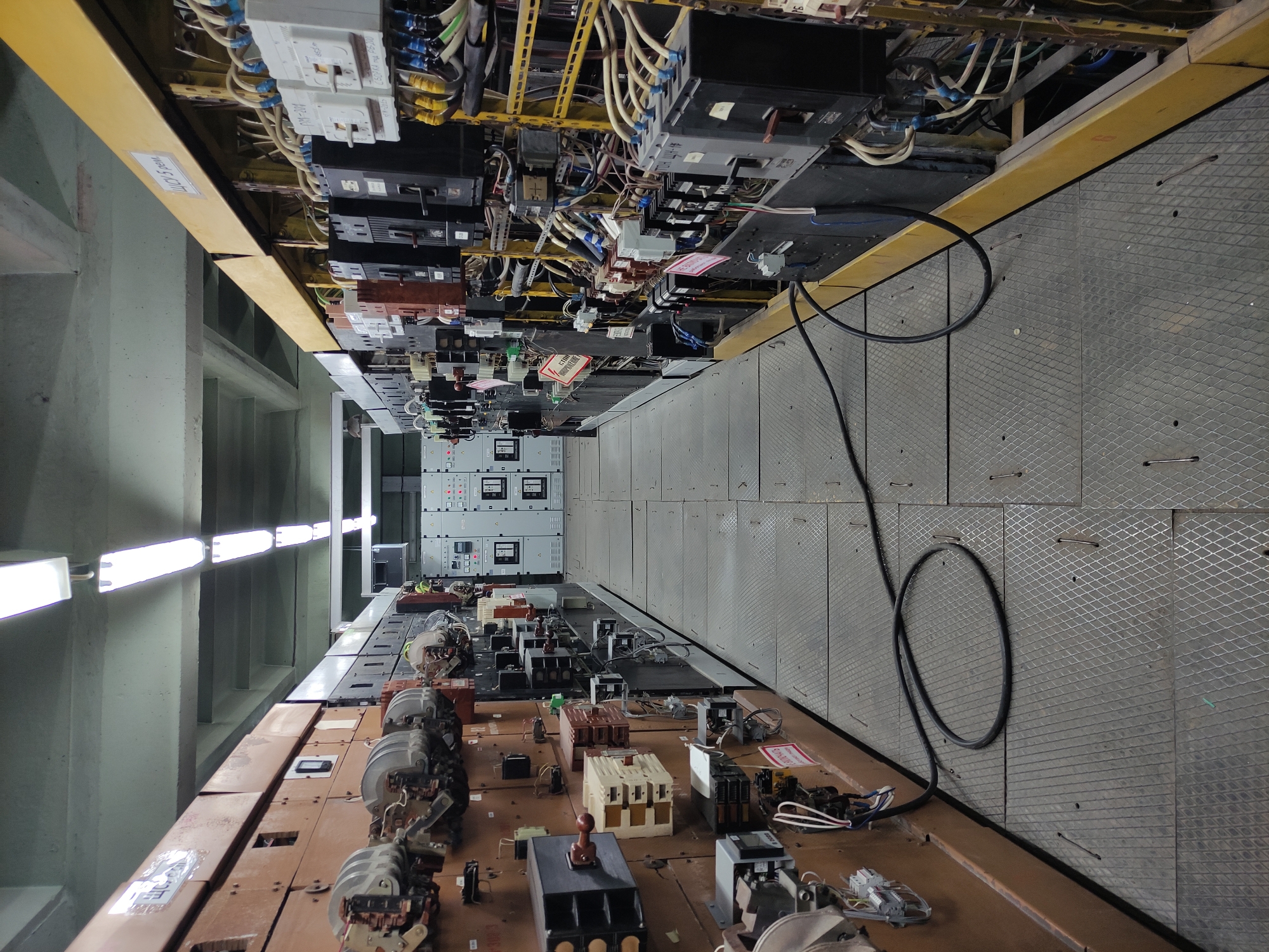 The image depicts the interior of an electrical substation. Cables, circuit breakers, and control panels are visible along the walls and above the floor. The walls are painted light green and illuminated by fluorescent lights. A black cable is visible on the floor. The floor itself is made of textured metal plating. No people are visible. While the image could potentially be from Pavlodar, Kazakhstan, based on similar infrastructure, it cannot be conclusively determined from visual elements alone.