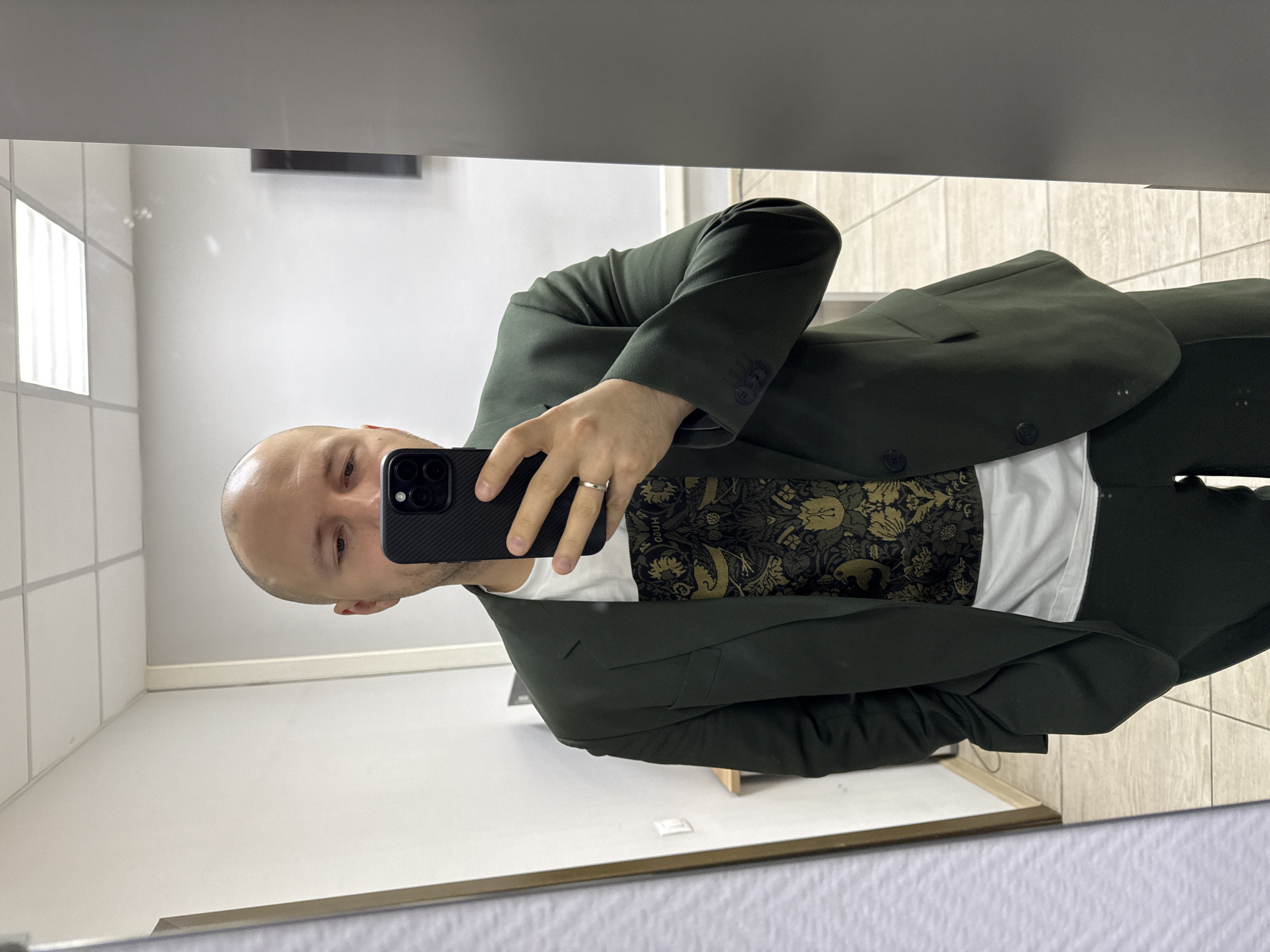 A bald man, appearing to be in his 30s or 40s, captures a mirror selfie in what seems to be an indoor setting in Krasnoyarsk, Russia. The man holds a black smartphone, likely an iPhone given its triple camera setup and case design, in his right hand, facing the mirror. He wears a dark green blazer and matching trousers, paired with a white t-shirt featuring a distinctive dark and gold ornate, possibly floral or baroque, pattern. The text "HUGO" is faintly visible as part of the shirt's pattern. A silver ring is visible on his left ring finger.
The setting appears to be a well-lit indoor space, possibly a dressing room or a public restroom, suggested by the white rectangular wall tiles on the left side of the reflection and the light-colored tiled floor. A rectangular light fixture is visible in the upper left of the mirror's reflection, and a dark, horizontal feature (perhaps a shelf or vent) is seen above. The man's expression is neutral as he poses for the picture. The image itself is rotated 90 degrees counter-clockwise.