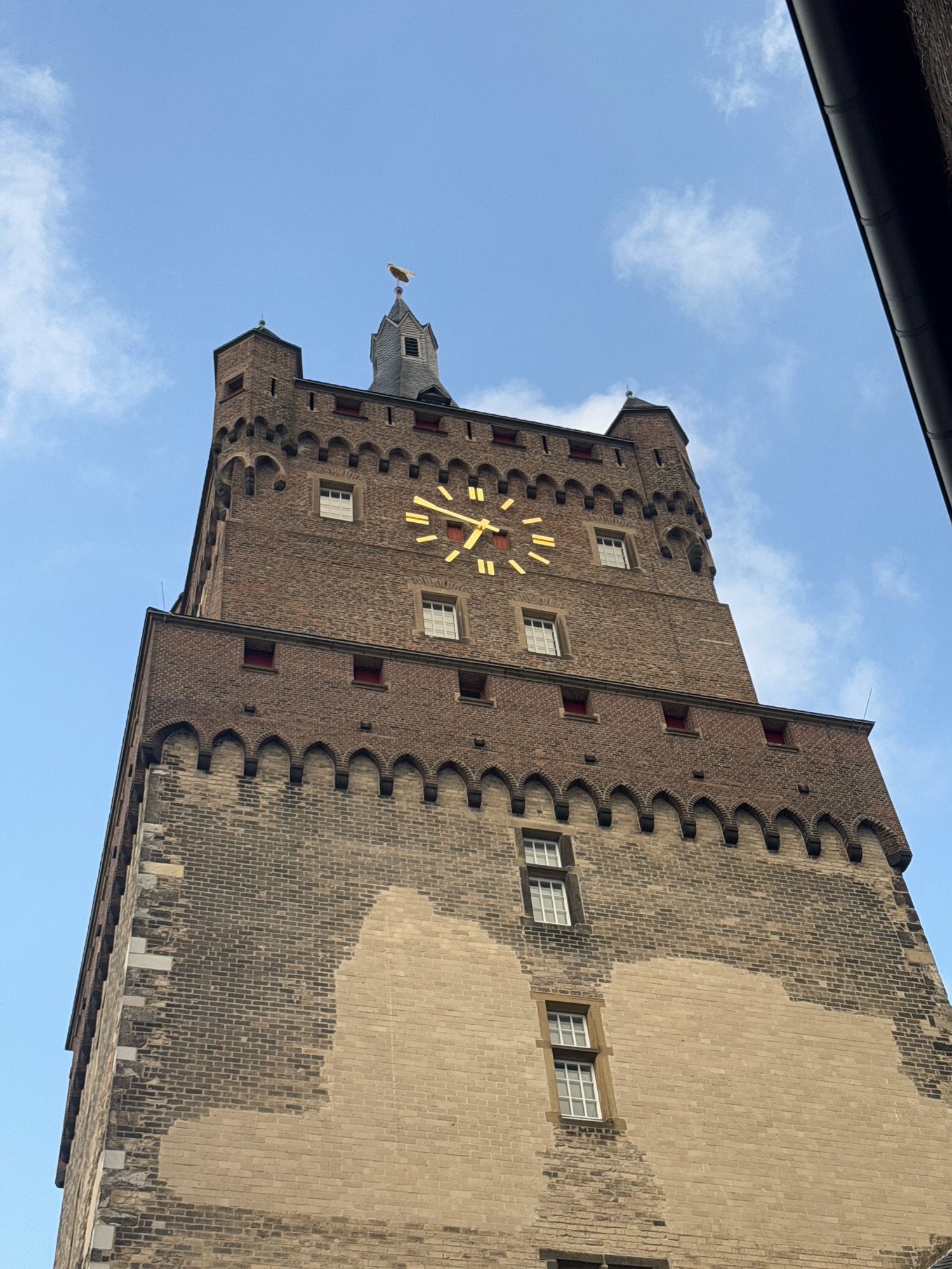 A low-angle, full shot captures the top portion of the historic Cleves Castle tower, known as the "Schwanenturm," against a bright blue sky with scattered white clouds. The tower, constructed from dark red and lighter beige brick, features crenellations and arched windows. A large, prominent golden clock face with Roman numerals is positioned midway up the tower. At the very top of the tower, a small spire is topped with a golden weather vane. The image is captured from a street-level perspective looking upward, emphasizing the tower's imposing height and architectural details. No people are visible in the frame.