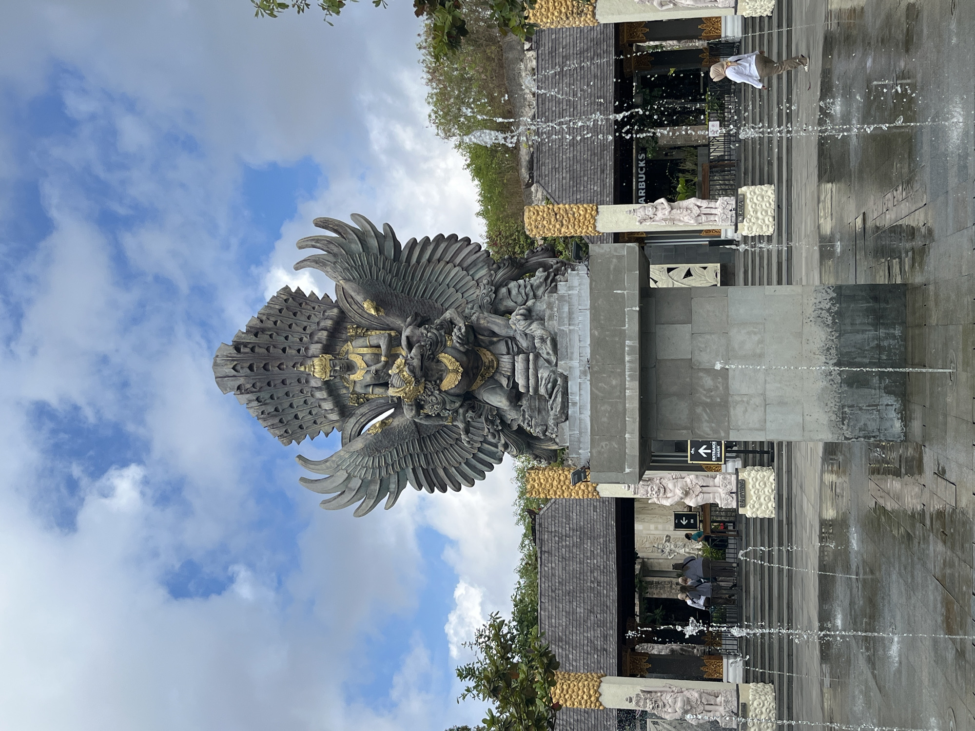The image captures a large, ornate statue of Garuda, a mythical bird-like creature, in front of a building with a thatched roof. The statue is dark gray with golden accents and depicts Garuda with a human-like torso and head, seated on its haunches with outstretched wings. In the background, a Starbucks is visible. The scene is set in a public area with fountains creating a water feature in the foreground. Several people are present, including a woman walking in the lower right corner and a group of people standing near the entrance of the building on the left. The sky above is partly cloudy. The location is identified as Garuda Wisnu Kencana Cultural Park in Ungasan, Kuta Selatan, Bali, Indonesia.
