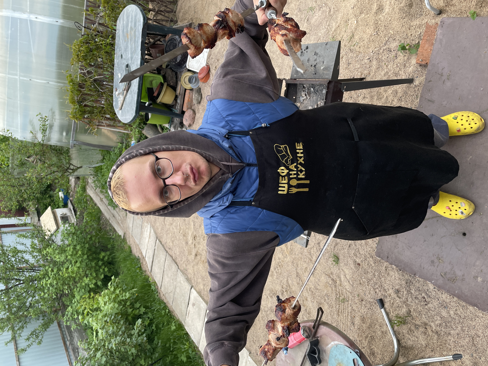 A person is cooking shish kebab outdoors in Sestroretsk, Russia. The individual is wearing a brown hooded sweatshirt, a blue vest, a black apron with gold lettering that reads "ШЕФ НА КУХНЕ" (Chef in the Kitchen), and yellow clogs. They are holding two metal skewers with cooked meat on them. In the background, there is a greenhouse, a green table, and a grill. The ground is covered with sand and patches of grass.