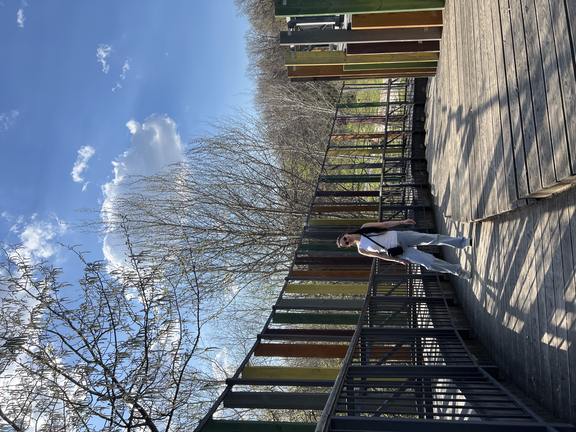 A full-length image depicting a woman standing on a multi-colored staircase structure. She's fair-skinned, with long dark hair, wearing a white top, light-wash jeans, white sneakers, sunglasses, and a black bag. She faces left in the frame.
The setting is an outdoor wooden platform. To the left of the woman is a metal staircase with steps painted in alternating colors: green, yellow, brown, and gray. In the background are trees with bare branches, suggesting late autumn or early spring. The sky is blue with fluffy white clouds.
Based on the visual characteristics, this location is likely located in Kazan, Russia.