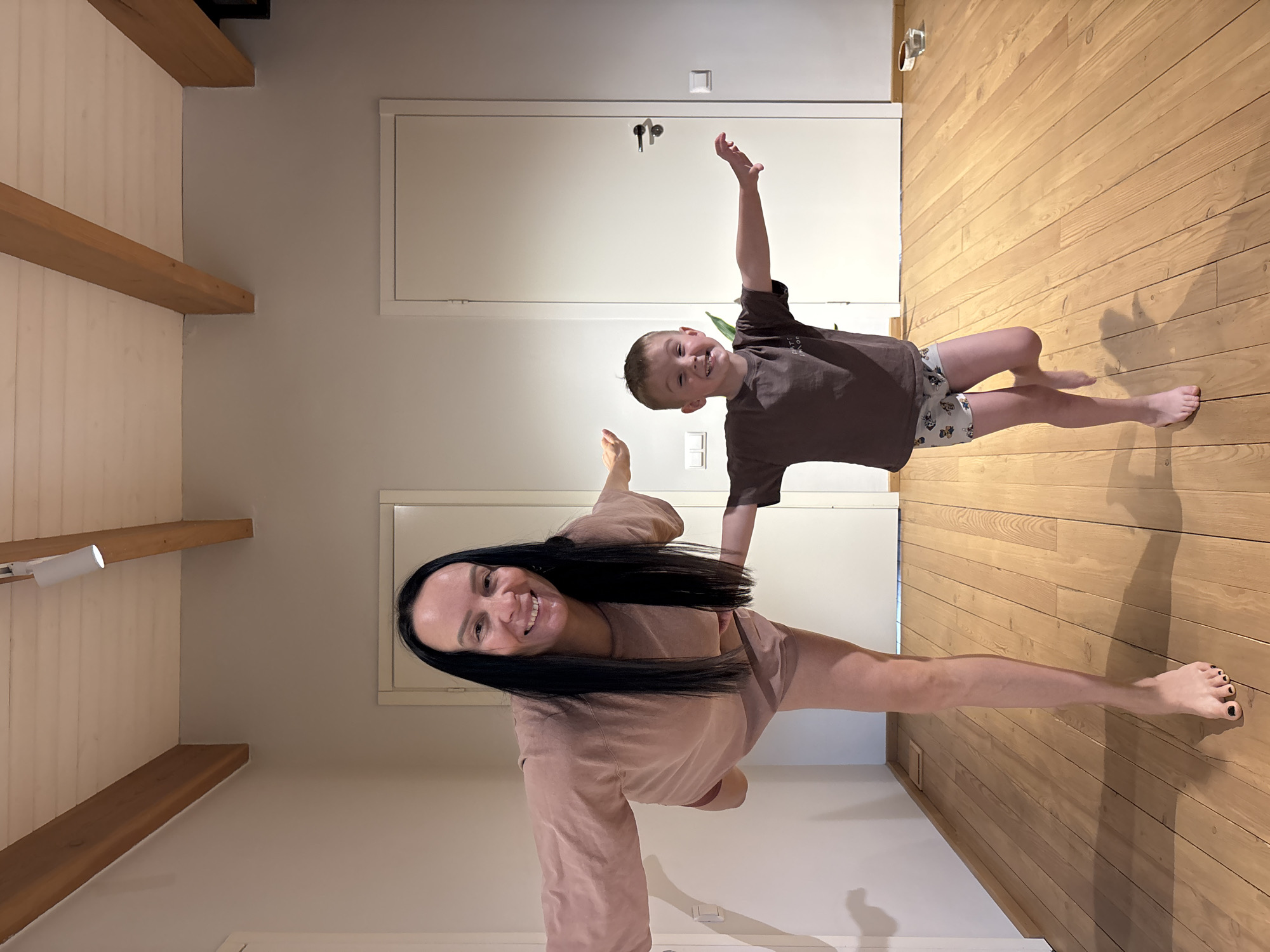 A woman and a young boy pose playfully in an interior space. The woman is dressed in a peach-colored loungewear set and has long dark hair. The boy wears a brown t-shirt and patterned shorts. Both have their arms raised. The interior features wooden floors, white walls, and wooden beams on the ceiling. There are two closed white doors in the background, one above the other. There is no confirmation of the location being in Chaikovsky, Russia.