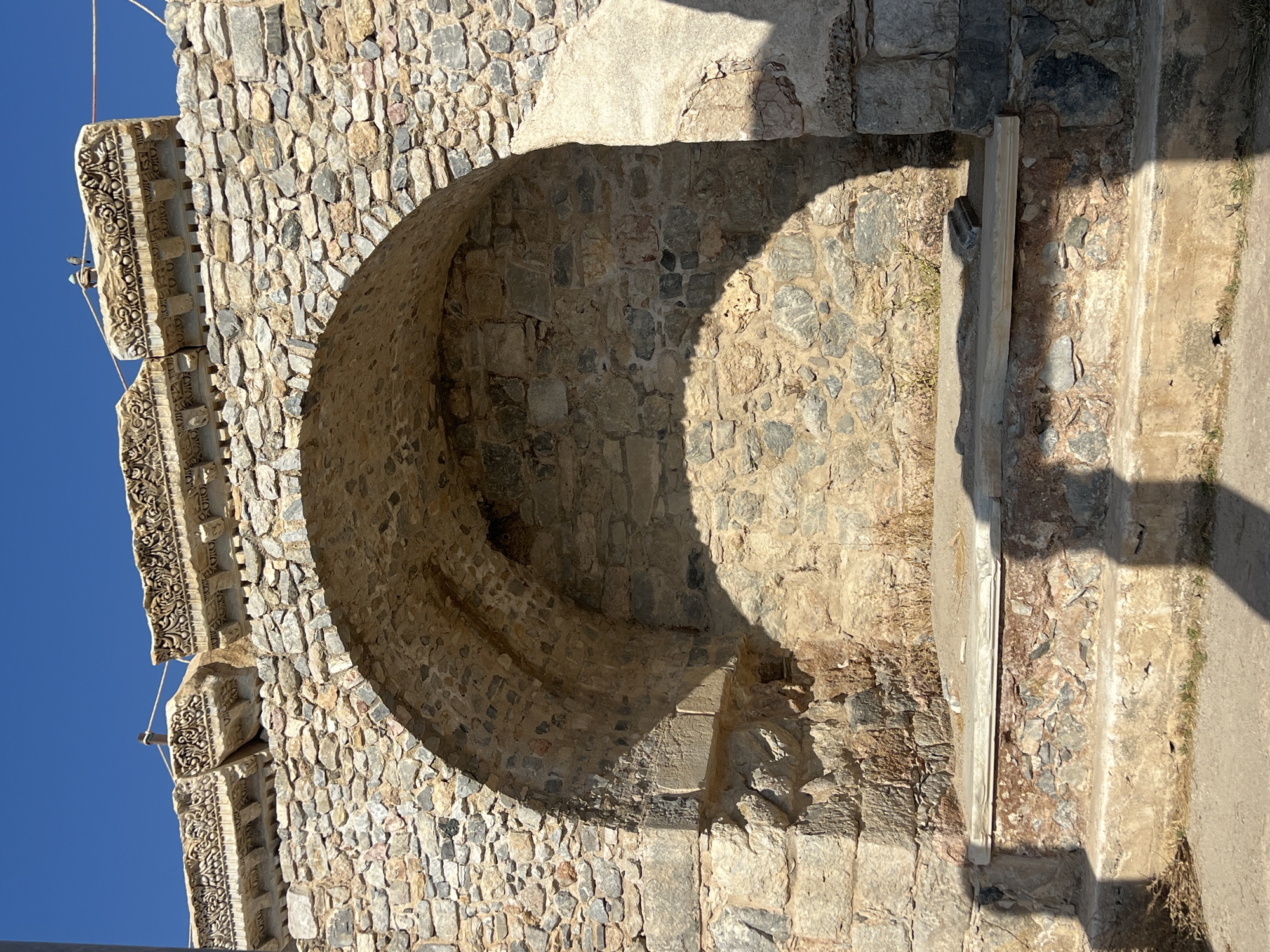 An arched alcove is formed by rough-hewn stone blocks and is set into a larger stone structure. The alcove contains a raised platform made of stone slabs, and the interior back wall is also constructed of stone. Above the arch, ornate carved stone friezes are visible, supported by a stone wall. The scene is bathed in sunlight, casting shadows that highlight the textures and contours of the stonework. The sky above is clear and blue.