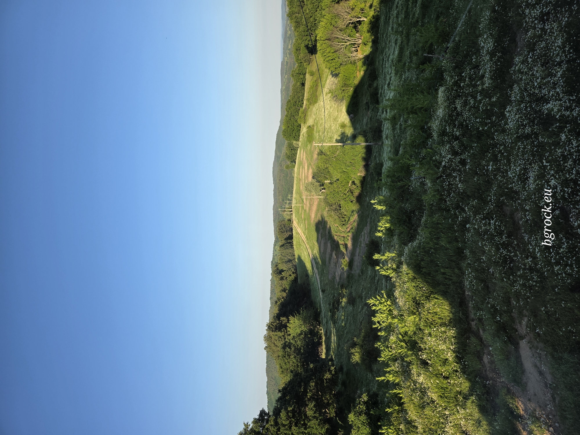 An outdoor scene captured at an angle, seemingly from an elevated position in or around the city of Chal, Bulgaria, shows an expanse of green, hilly landscape under a clear blue sky. A path or road traverses the hillside, partly obscured by vegetation. Utility poles and lines are visible, running alongside a lighter-colored strip of land cutting through the green. The vegetation is dense, possibly composed of grass, foliage, and some flowering plants. The watermark "bgrock.eu" is present in the lower right of the image.